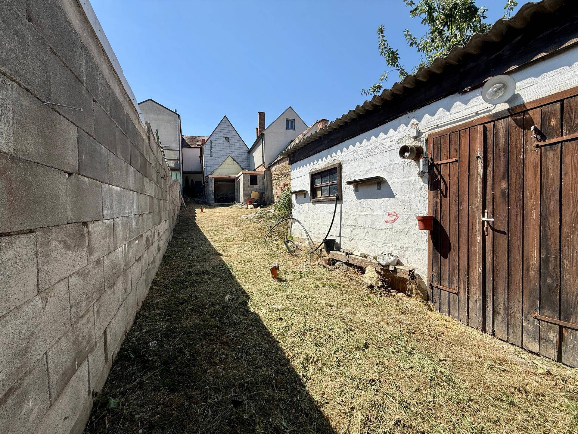 Blick in einen ländlichen Hinterhof mit alten Gebäuden, Holztüren und einer hohen Betonmauer.