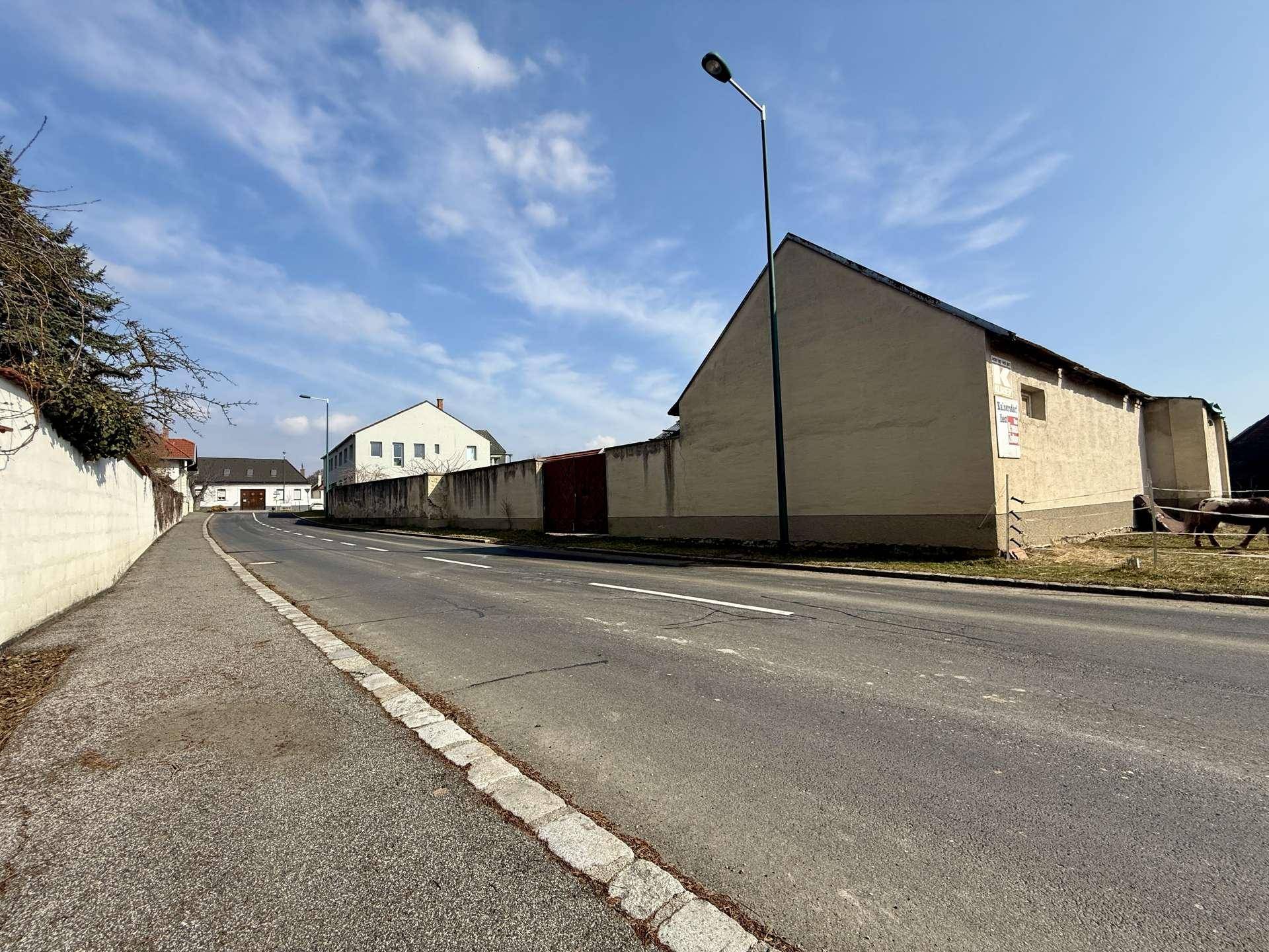 Ländliche Straße mit Gebäuden und einem langen Zaun unter blauem Himmel.