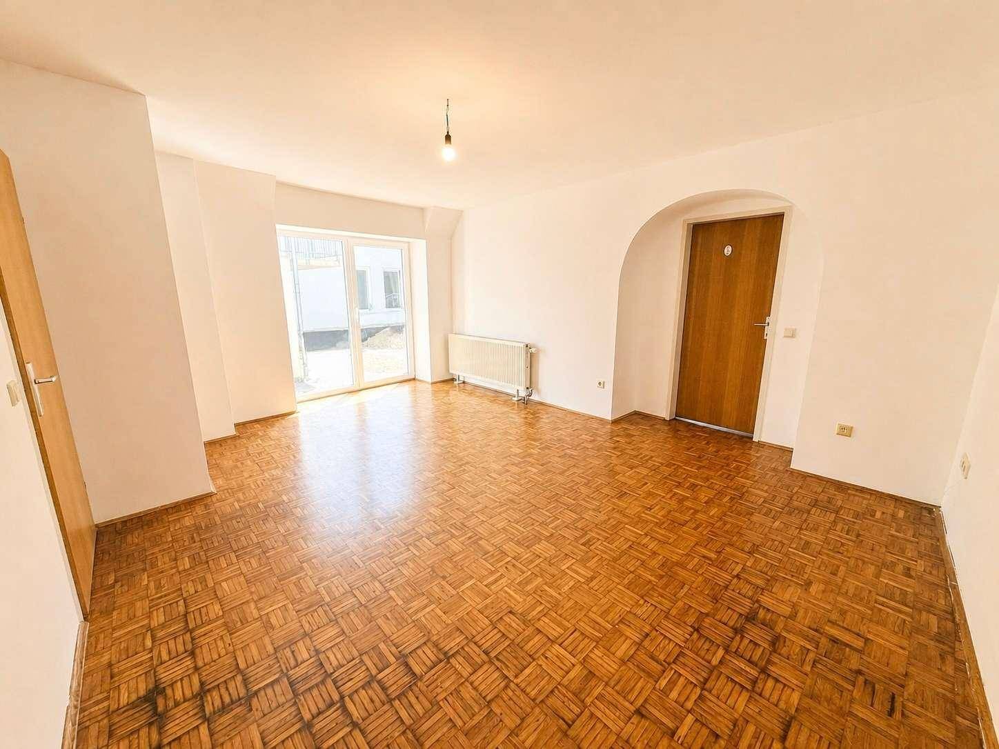 Helles Wohnzimmer mit Fischgrätparkettboden, weißen Wänden und Zugang zu einem Außenbereich durch eine große Glastür.