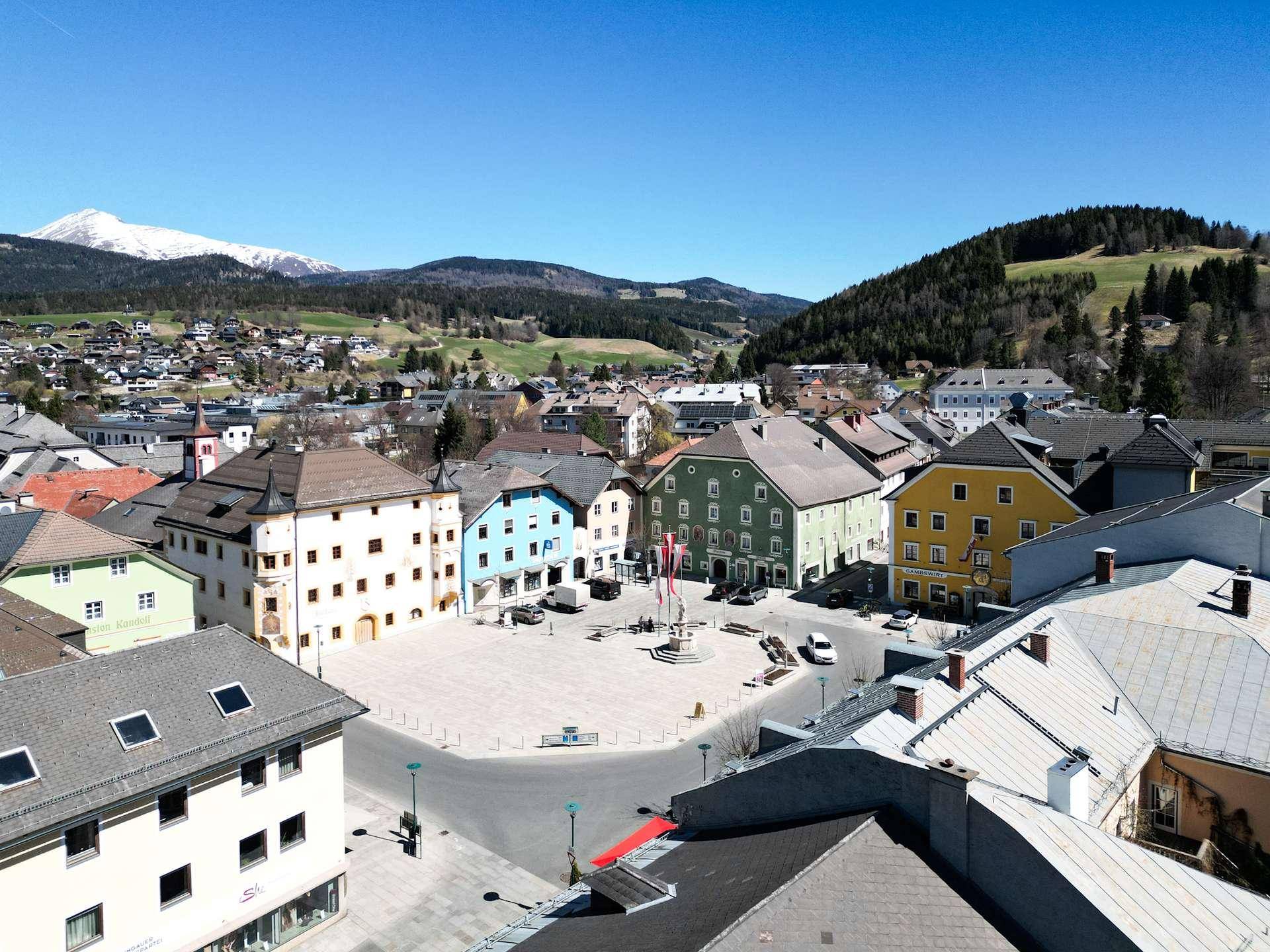 Weitläufiger Blick auf einen sonnigen Stadtplatz mit farbenfrohen Gebäuden und Bergen im Hintergrund.