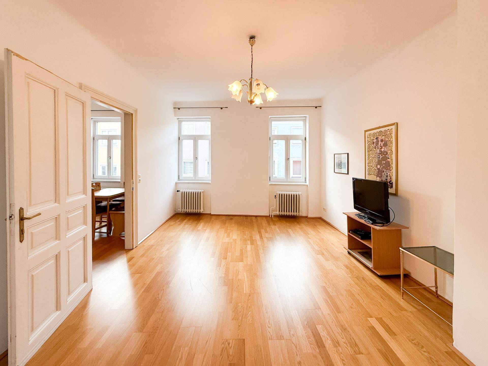 Unmöbliertes Wohnzimmer mit Holzboden, hohen Decken und Zugang zu einem weiteren Raum.