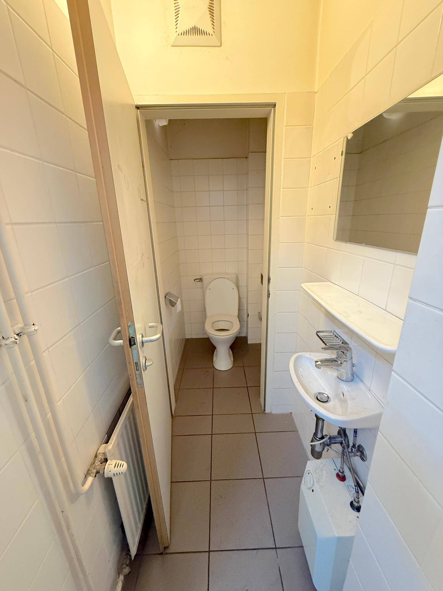 Einfache Toilette mit weiß gefliesten Wänden, kleinem Waschbecken und Heizkörper.