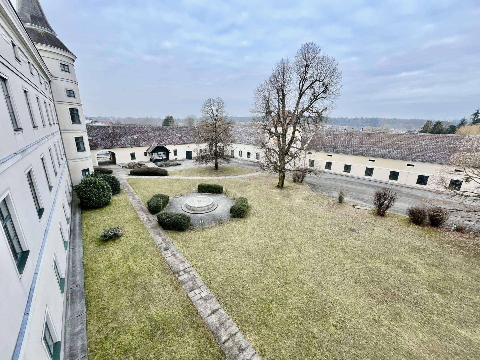 Großer Innenhof eines historischen Gebäudes mit Rasenfläche, Bäumen und einem Brunnen.