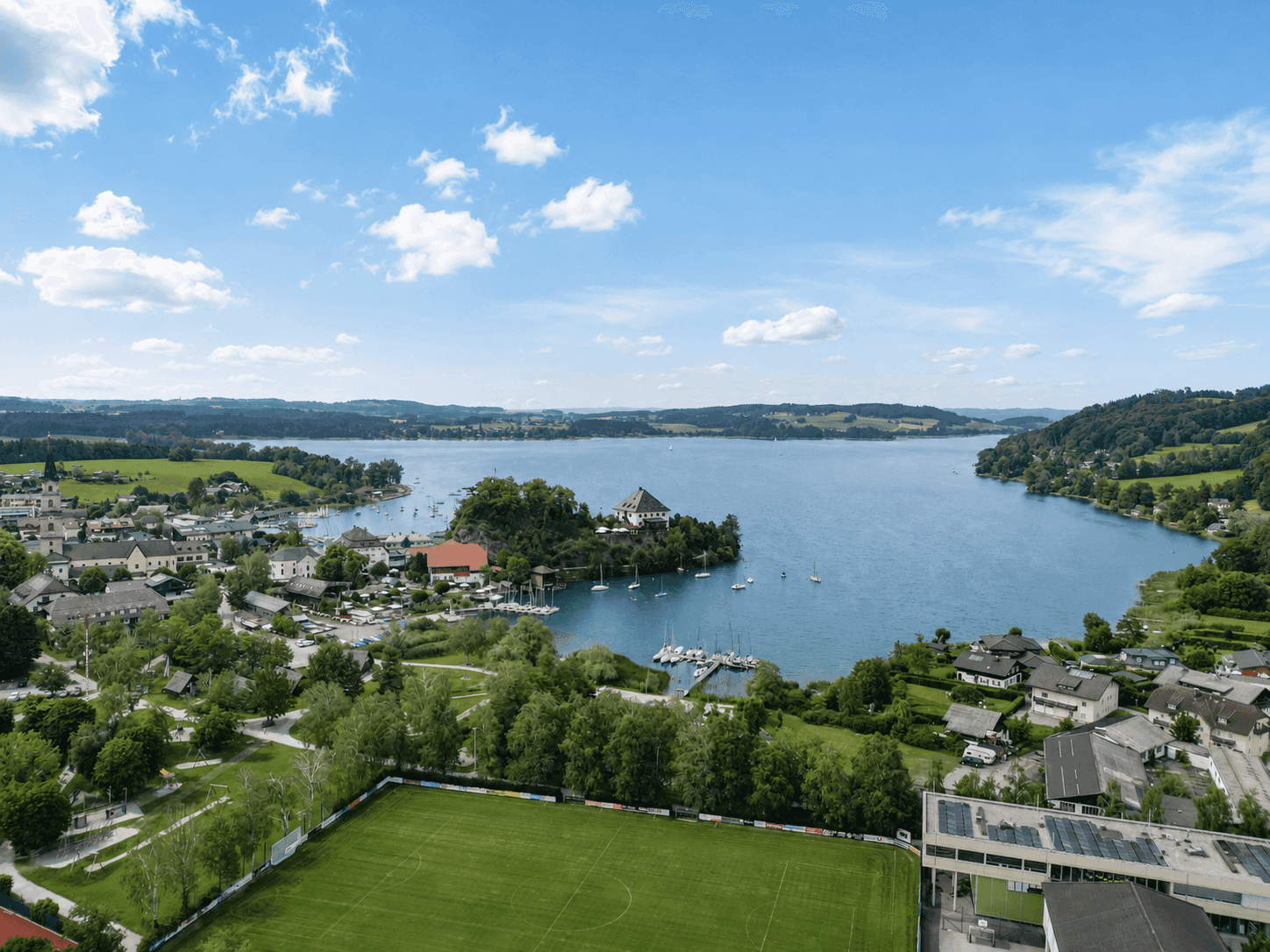Luftaufnahme einer malerischen Seenlandschaft mit angrenzender Ortschaft und Sportplatz unter blauem Himmel.