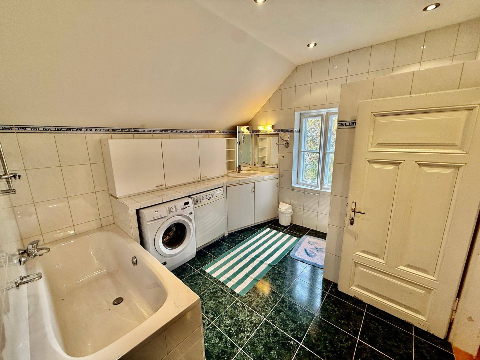 Badezimmer mit Badewanne, Waschmaschine und grünen Fliesen, ausgestattet mit Spiegelschrank.