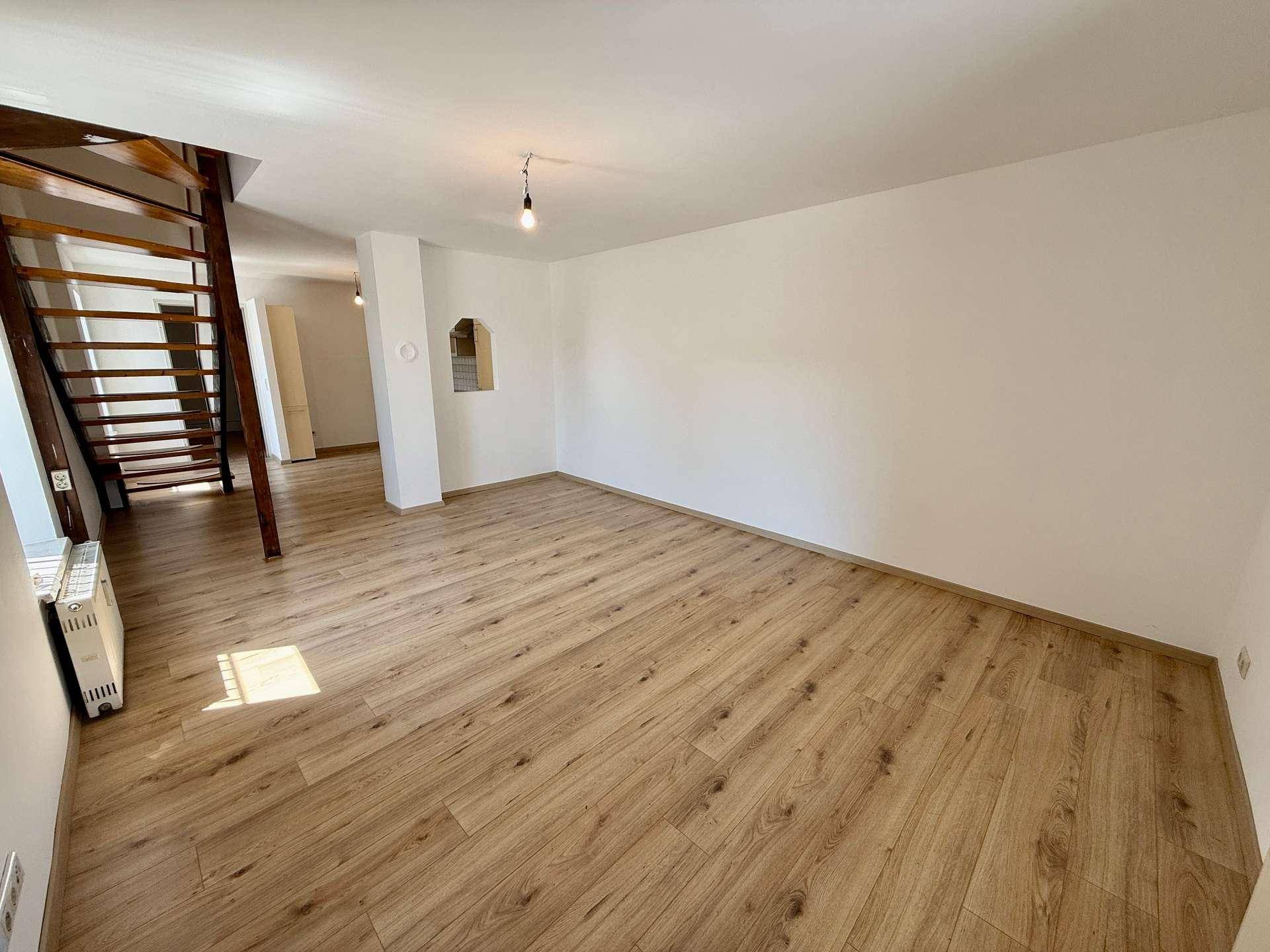 Geräumiges, leeres Wohnzimmer mit hellem Laminatboden und Zugang zur Holztreppe.