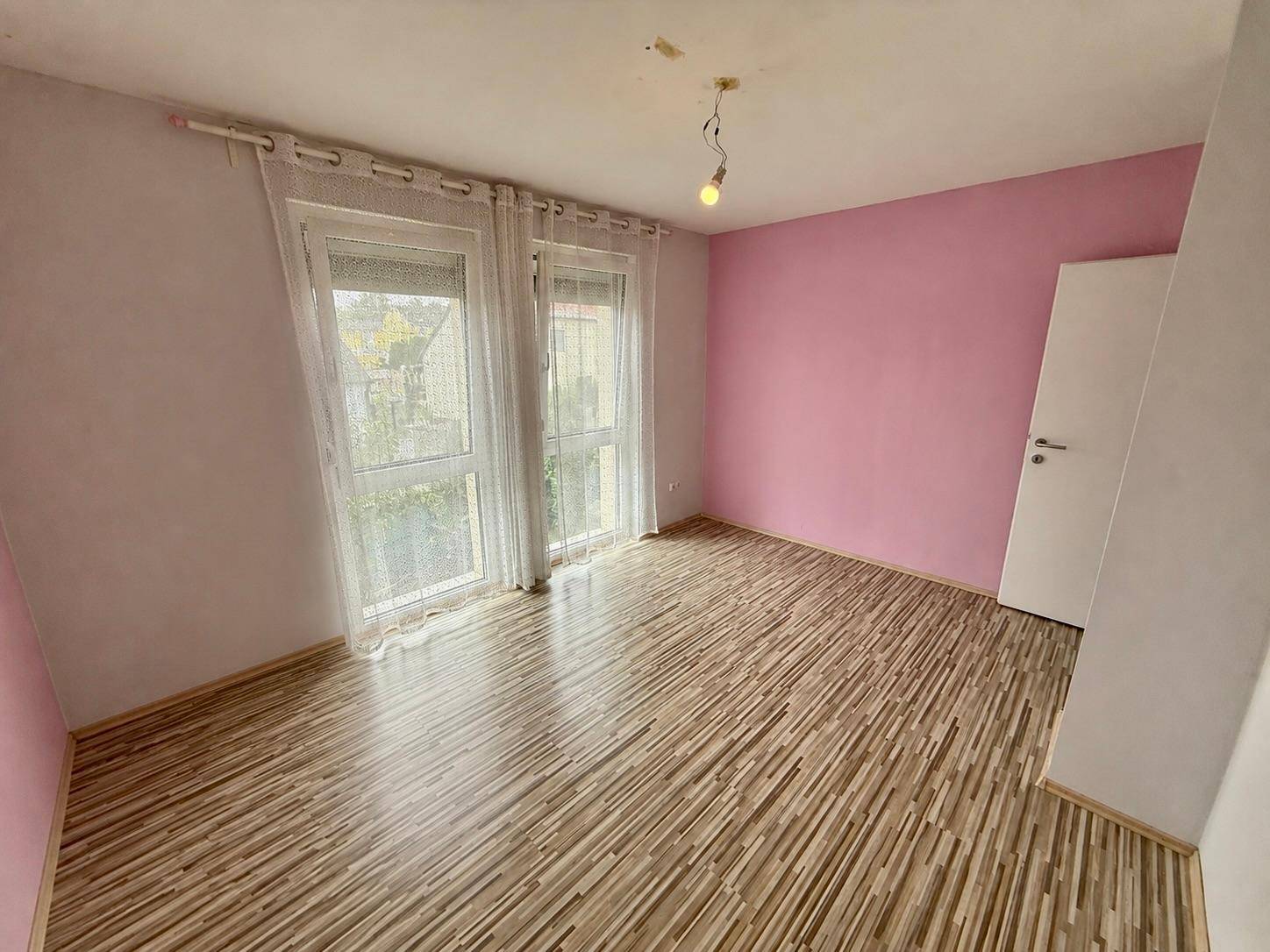 Freundliches Zimmer mit rosa Wänden, Laminatboden und zwei Fenstern.