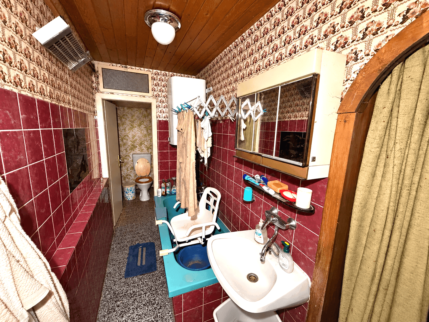 Badezimmer mit roten Fliesen, kleiner Badewanne und Waschbecken, ältere Ausstattung.