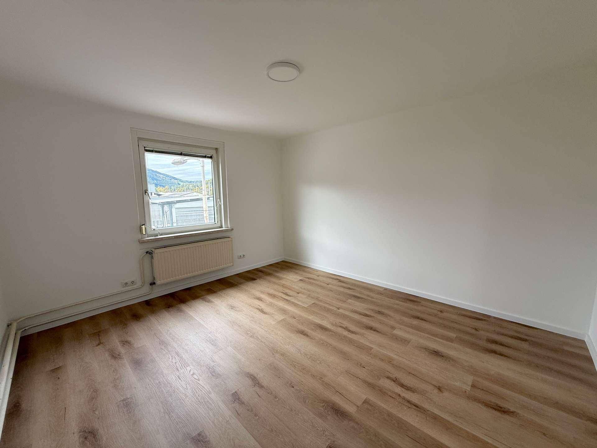 Helles Zimmer mit Laminatboden, weißen Wänden und einem Fenster mit Blick ins Grüne.