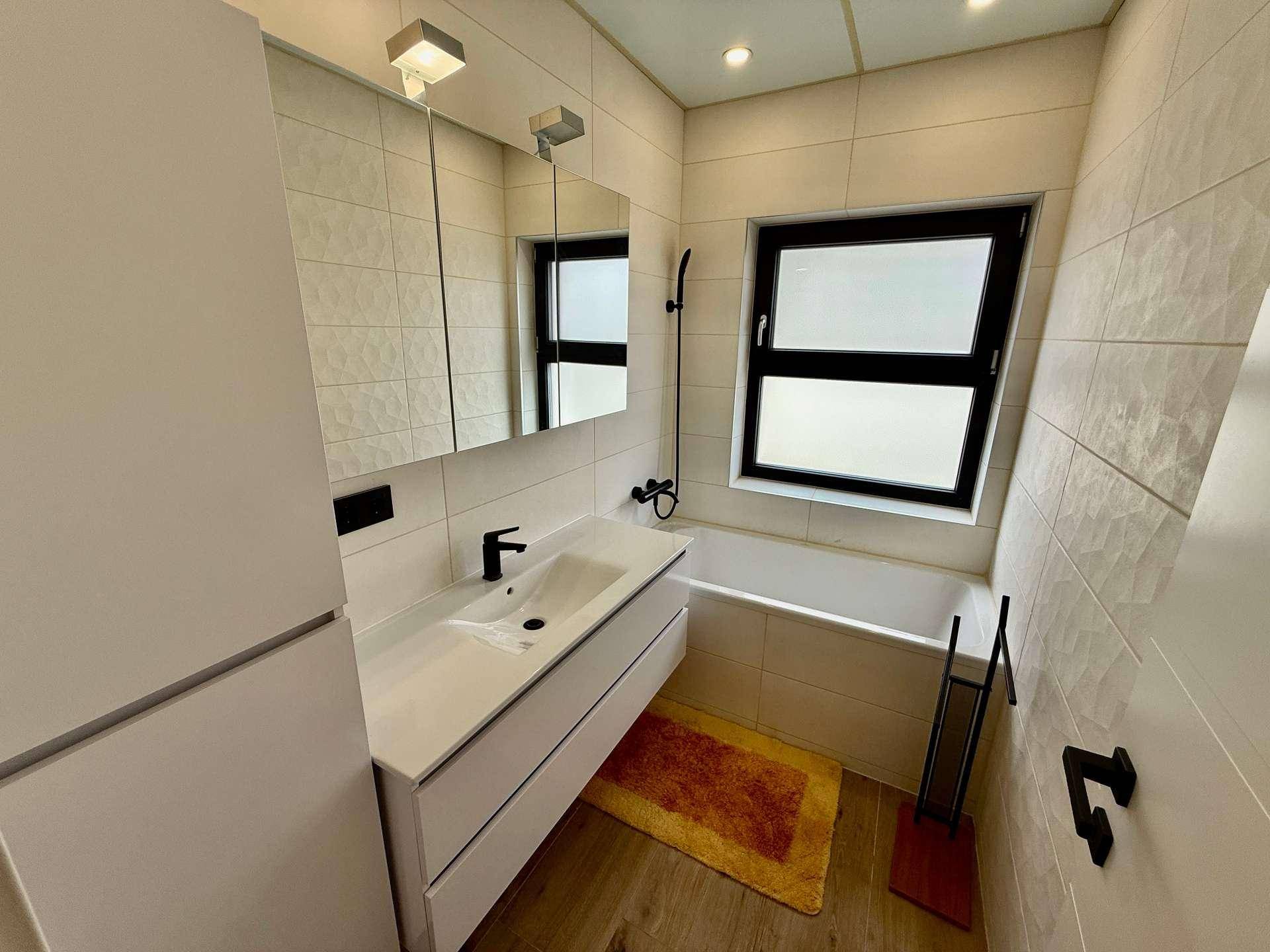 Modernes Badezimmer mit Badewanne, Waschtisch und Fenster, ausgestattet mit weißen Fliesen.