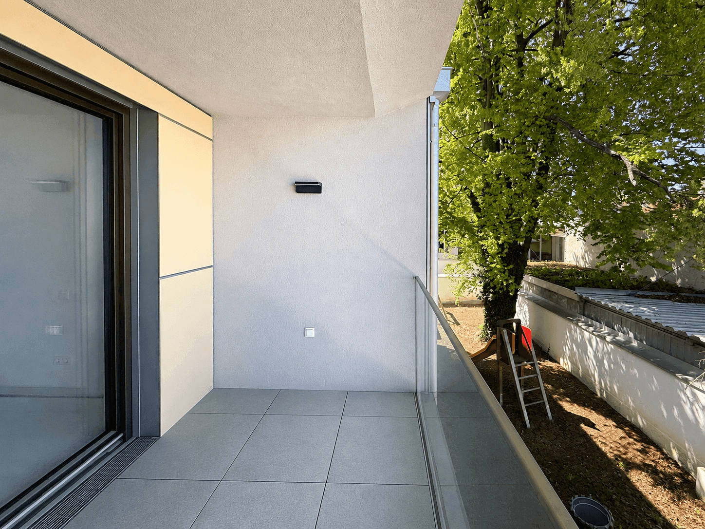 Heller Balkon mit Fliesen und Blick auf einen Baum sowie einen Spielplatz.