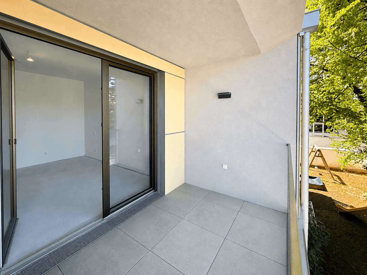 Moderner Balkon mit Fliesenboden und Glasgeländer, angrenzend an einen Baum.