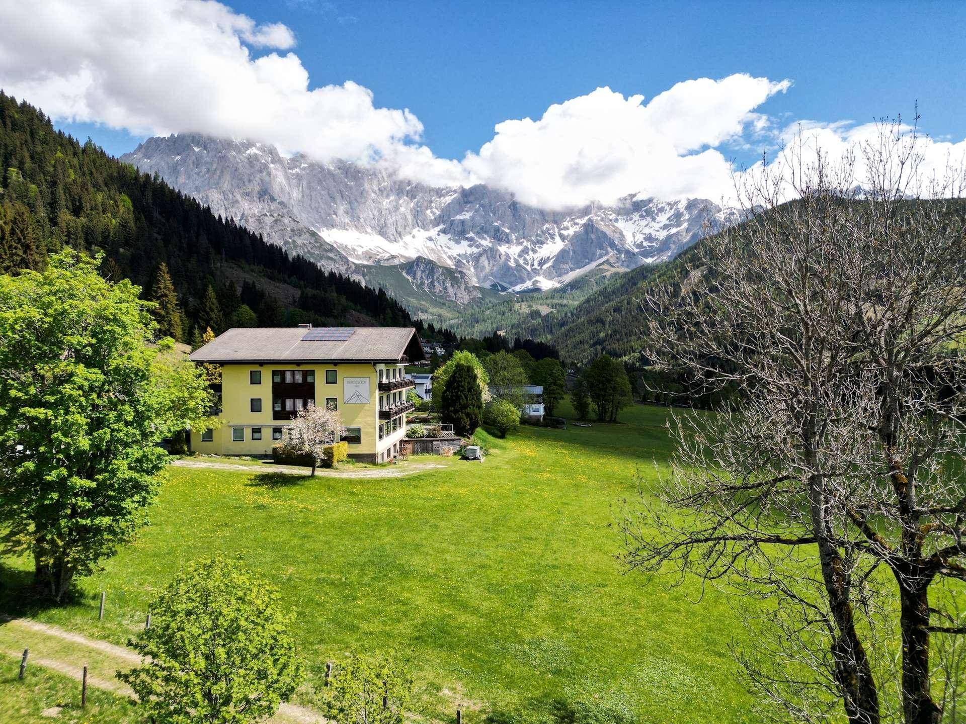 Panoramablick auf ein Gebäude inmitten weitläufiger grüner Wiesen mit majestätischen Bergen im Hintergrund.