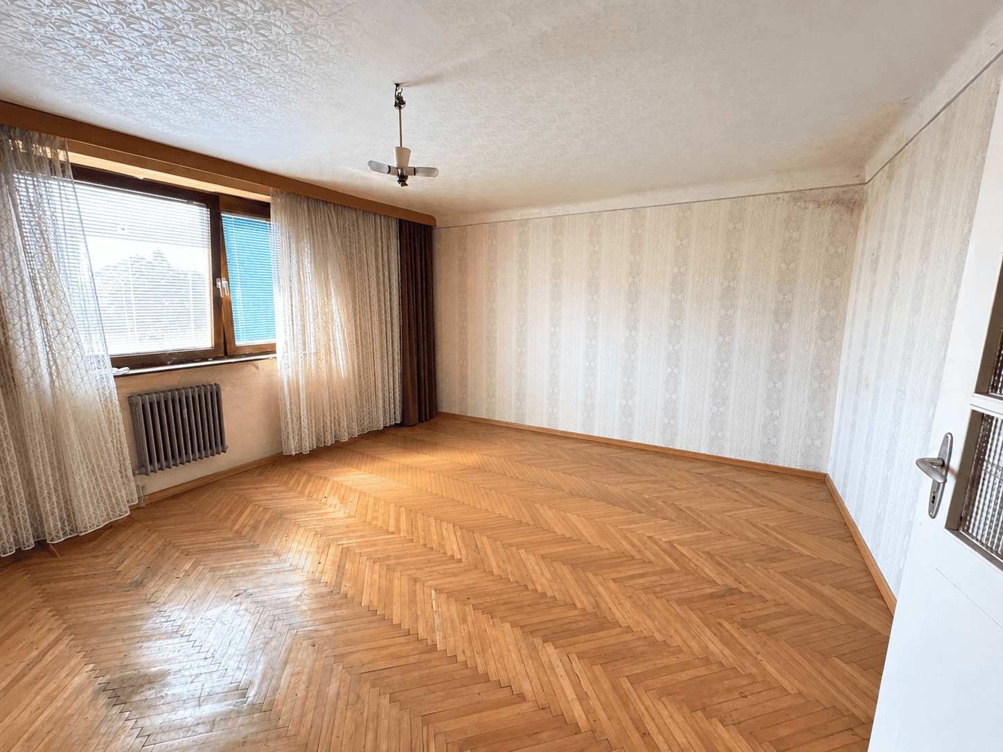 Geräumiges Zimmer mit Fischgrätparkett, gemusterter Tapete und einem Fenster mit Jalousien.