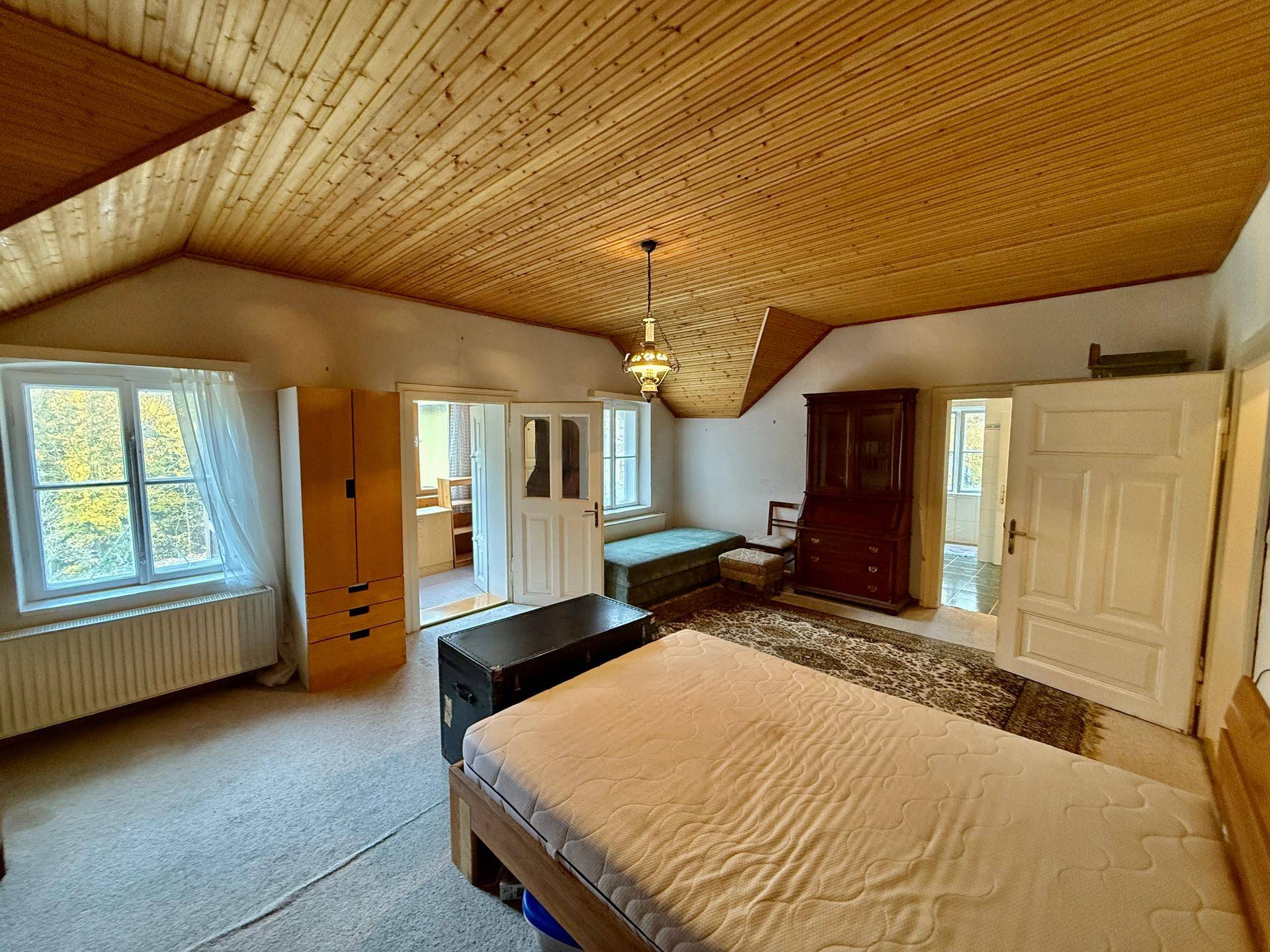 Schlafzimmer mit schräger Holzdecke, Teppichboden und Blick ins Grüne durch die Fenster.