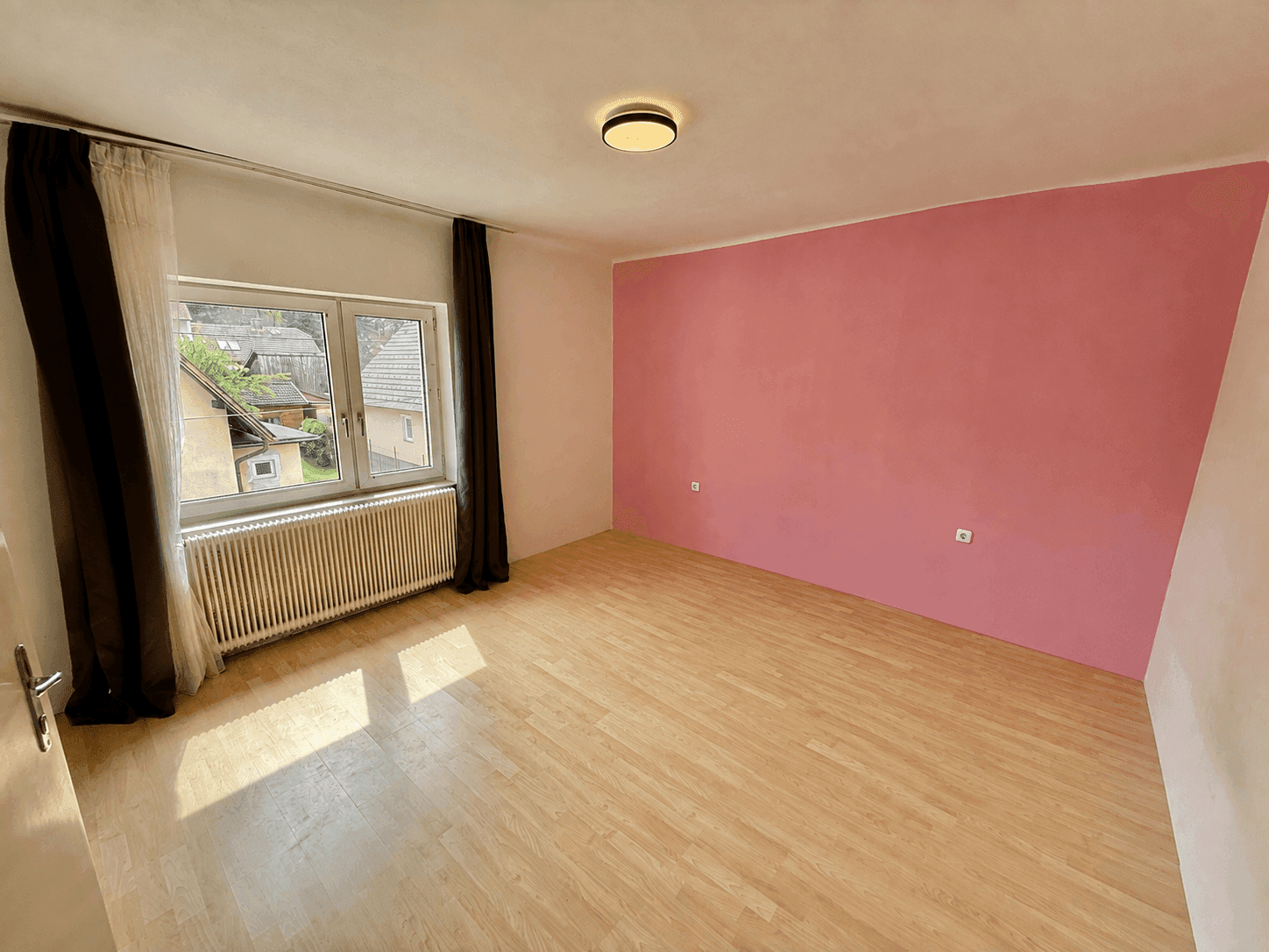 Helles Schlafzimmer mit Holzboden und einer rosa Akzentwand, gut beleuchtet durch ein großes Fenster.