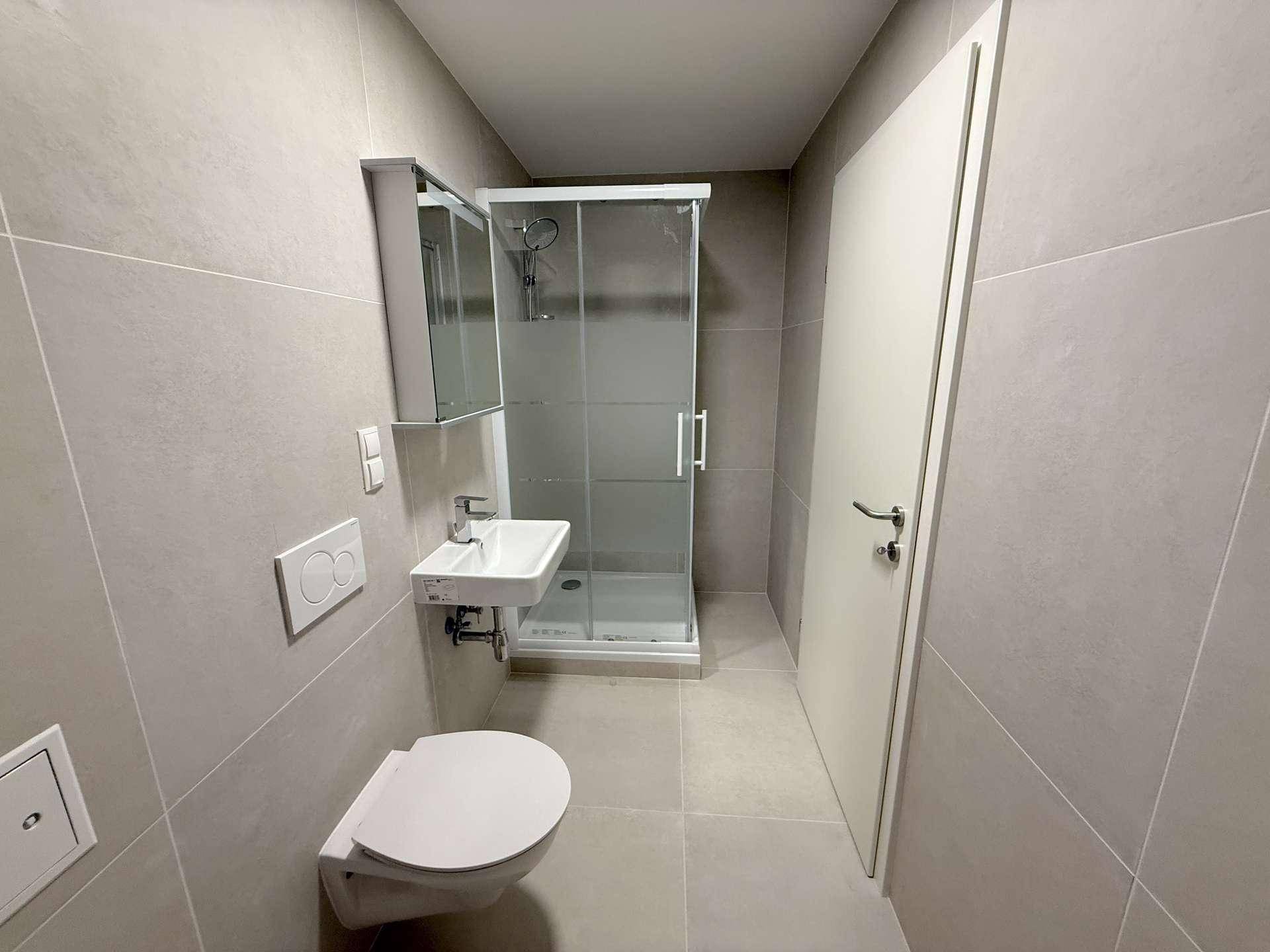 Modernes Badezimmer mit grauen Fliesen, wandhängendem WC, kleinem Waschbecken und Duschkabine.