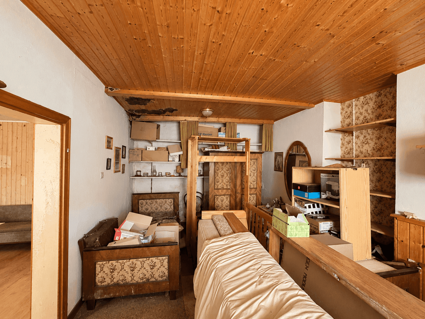 Schlafzimmer mit Holzdecke und Tapeten, derzeit als Abstellraum genutzt.