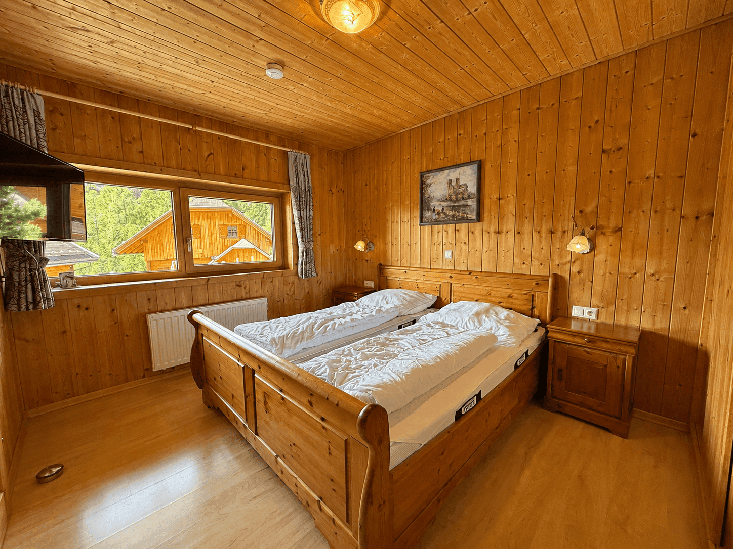 Schlafzimmer mit Doppelbett aus Holz, Holzvertäfelung an Wänden und Decke, mit Blick ins Grüne.
