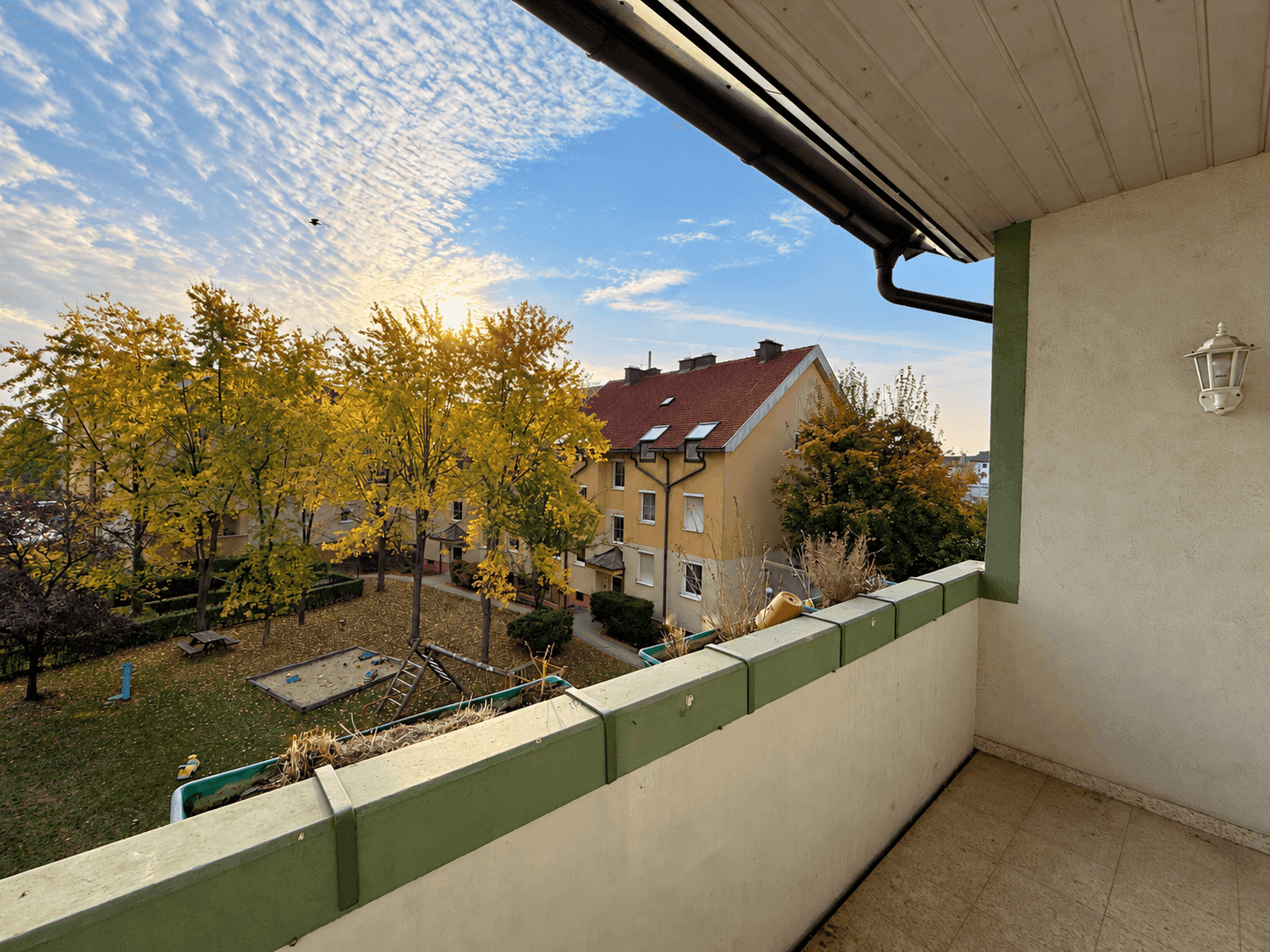 Balkon mit Blick auf einen grünen Innenhof und Spielplatz bei Sonnenuntergang.