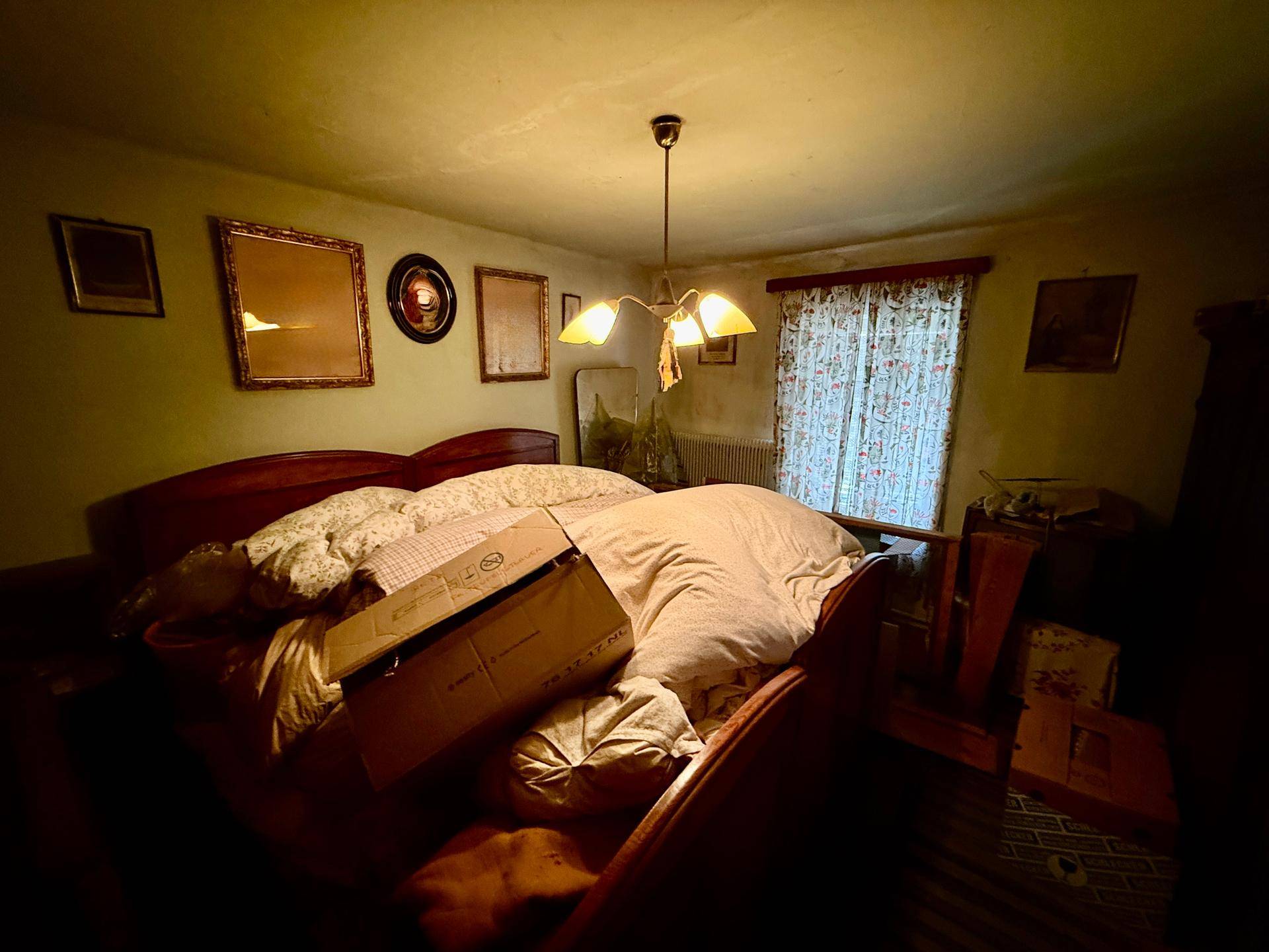Dunkles Schlafzimmer mit großem Bett, traditionellen Bildern an der Wand und Vorhängen am Fenster.