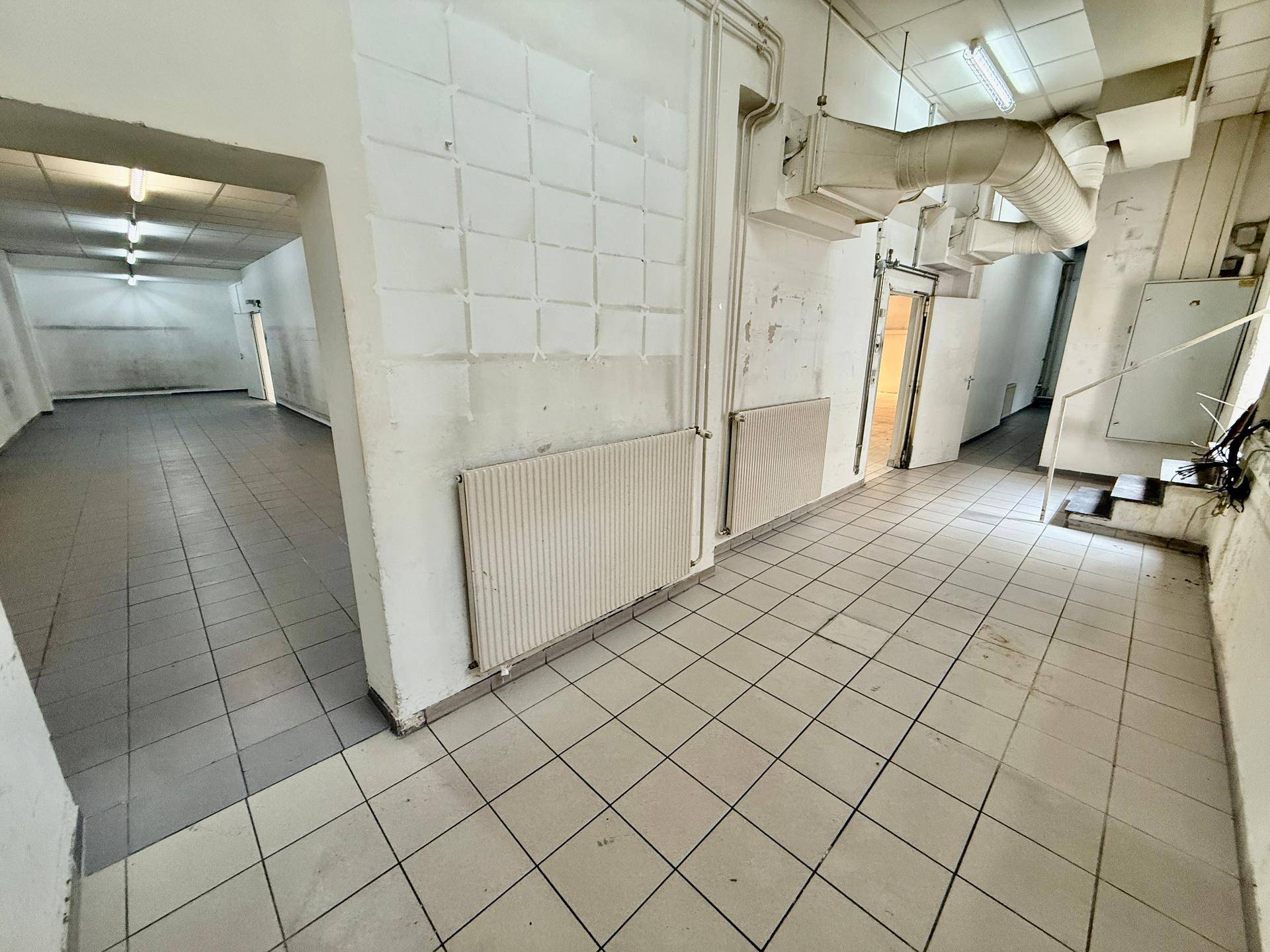 Leere Gewerbefläche mit grauen Fliesen, sichtbaren Heizkörpern und freiliegenden Lüftungsrohren an der Decke.