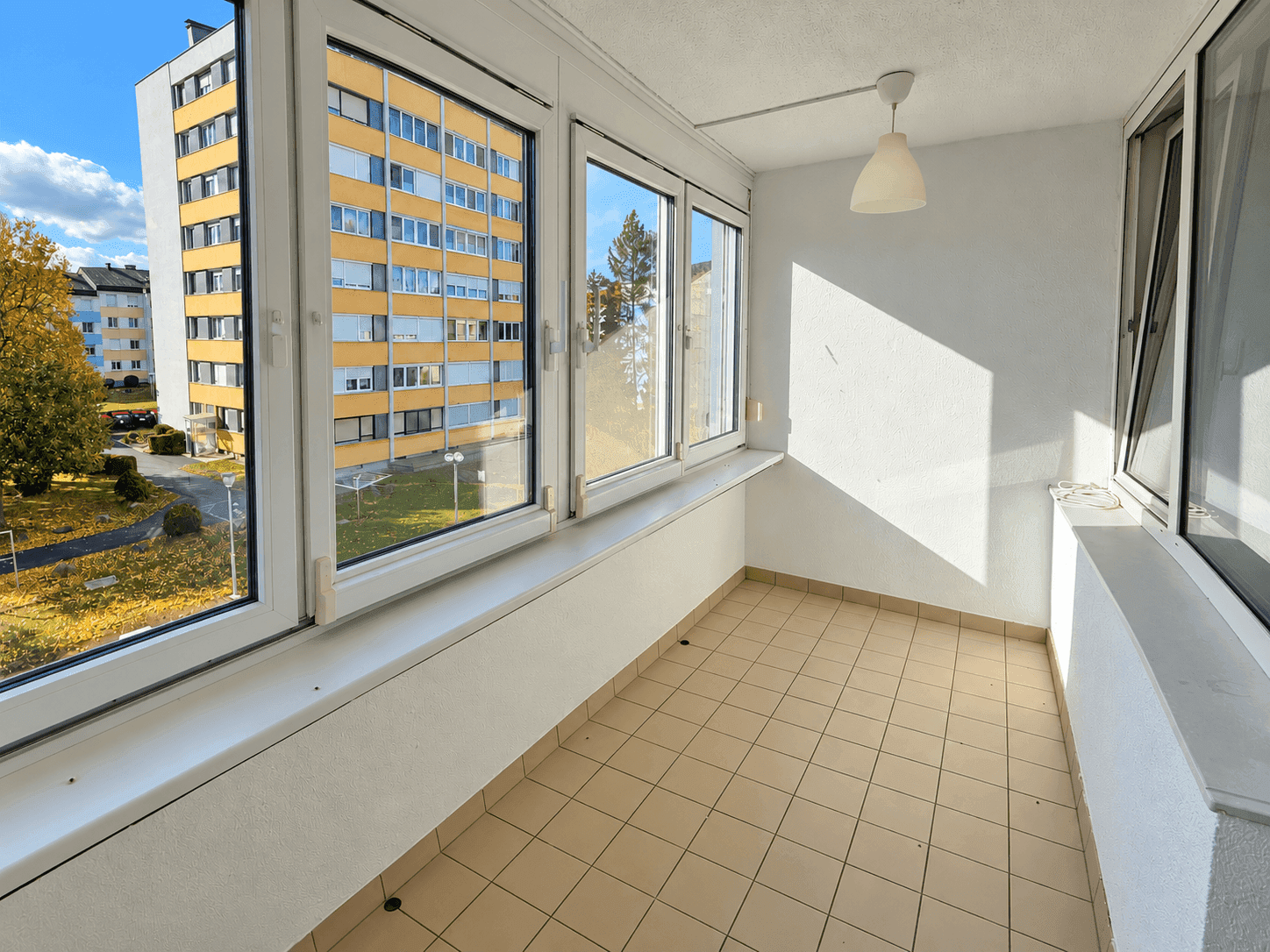Verglaster Balkon mit Fliesenboden und weitem Blick auf die Wohnanlage und die herbstliche Natur.