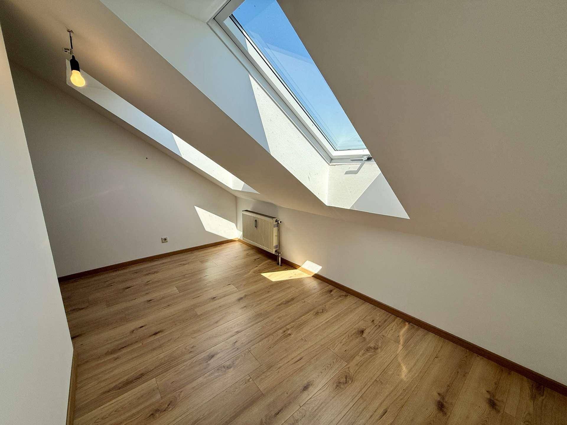Helles Dachzimmer mit schrägen Wänden, Laminatboden und zwei Dachfenstern.