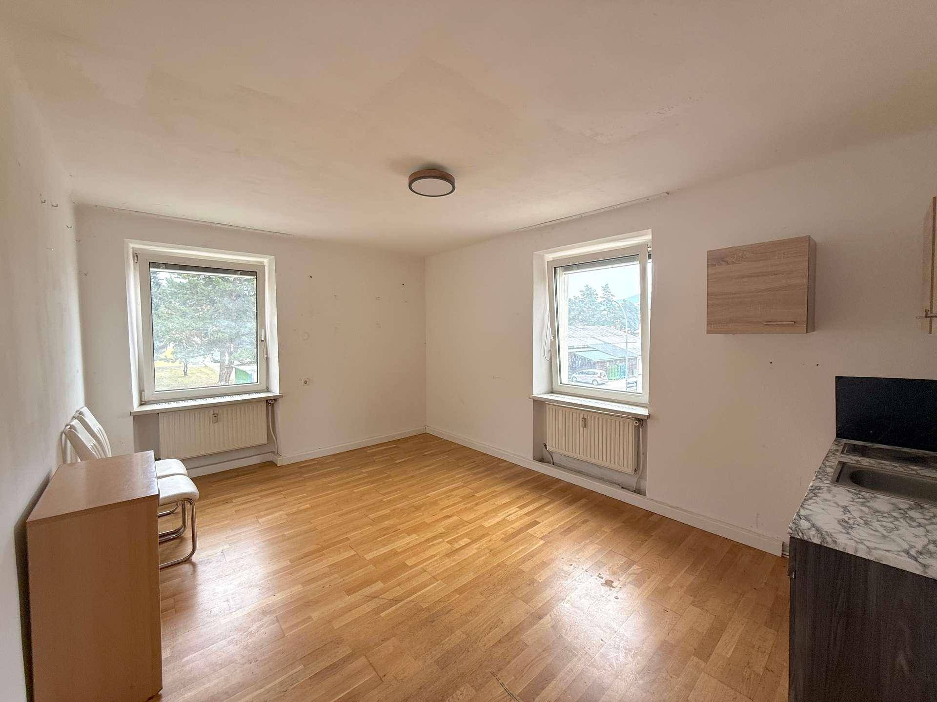 Geräumiges Wohnzimmer mit zwei Fenstern, Parkettboden und einfacher Küchenzeile.