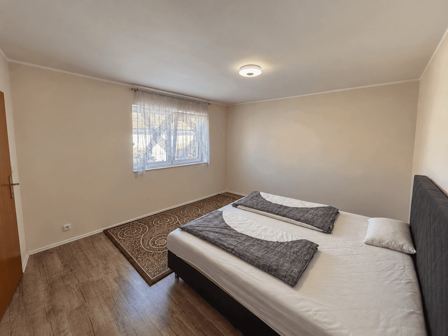 Helles Schlafzimmer mit Doppelbett, Laminatboden und Fenster mit Gardinen.