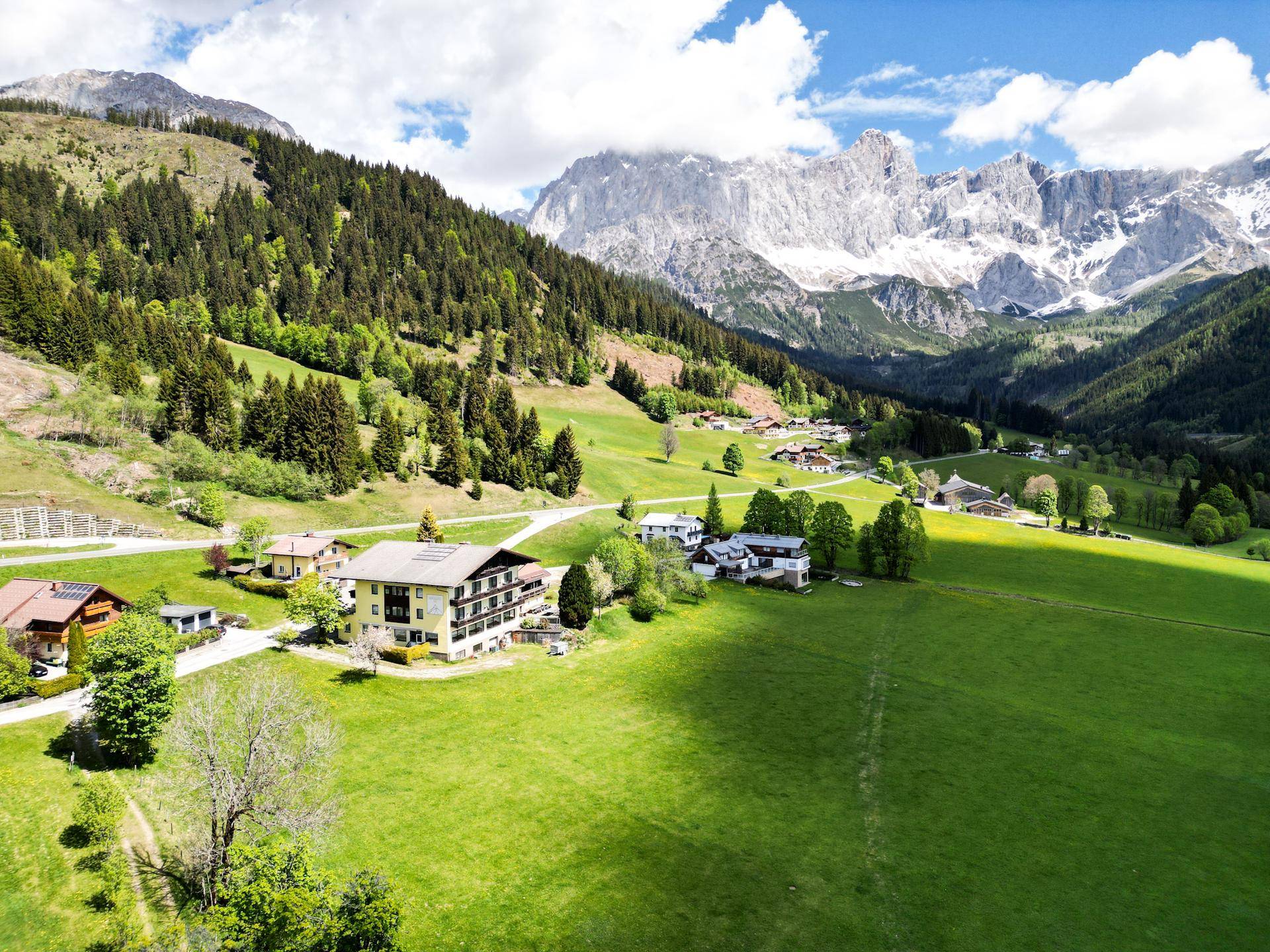 Weitläufige Ansicht eines gelben Gebäudes in einem Tal, umgeben von Wäldern, Wiesen und schneebedeckten Bergen.