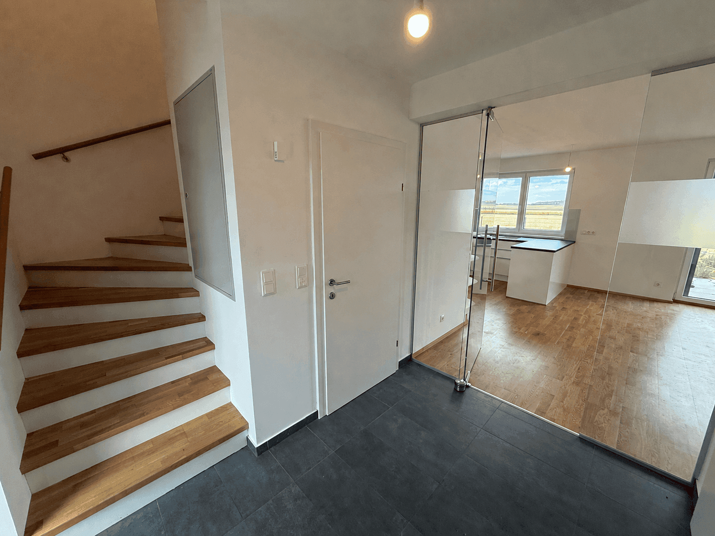 Heller Eingangsbereich mit Holztreppe und Blick durch eine Glastür in den offenen Wohnbereich.