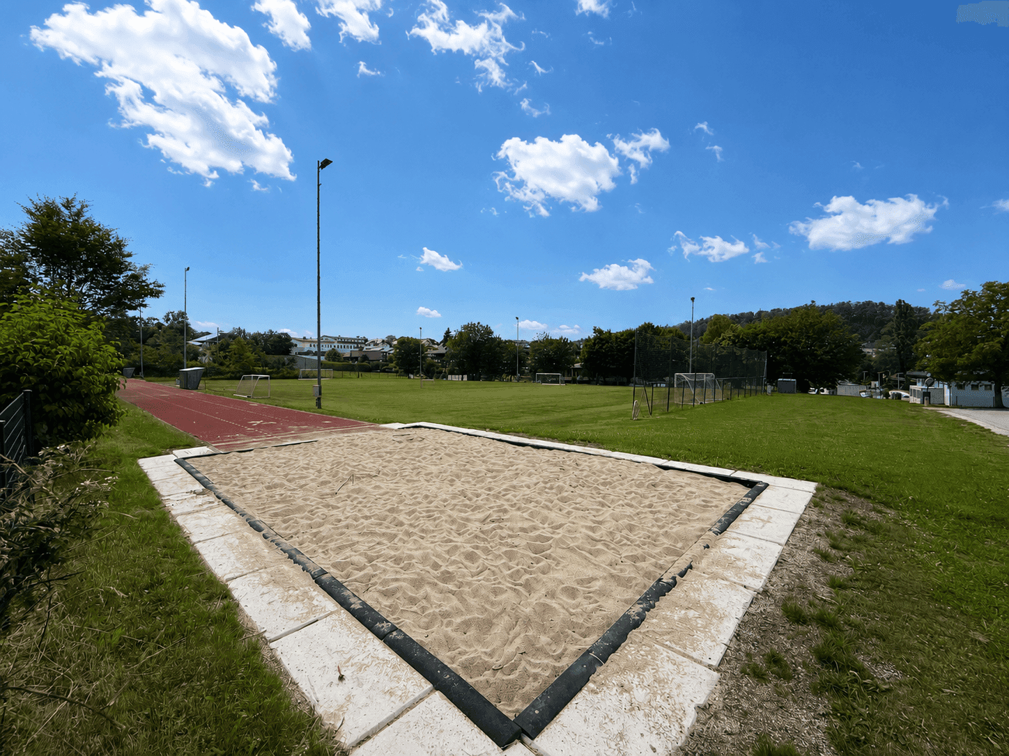 Sportplatz mit Weitsprunganlage und Rasenflächen unter blauem Himmel.