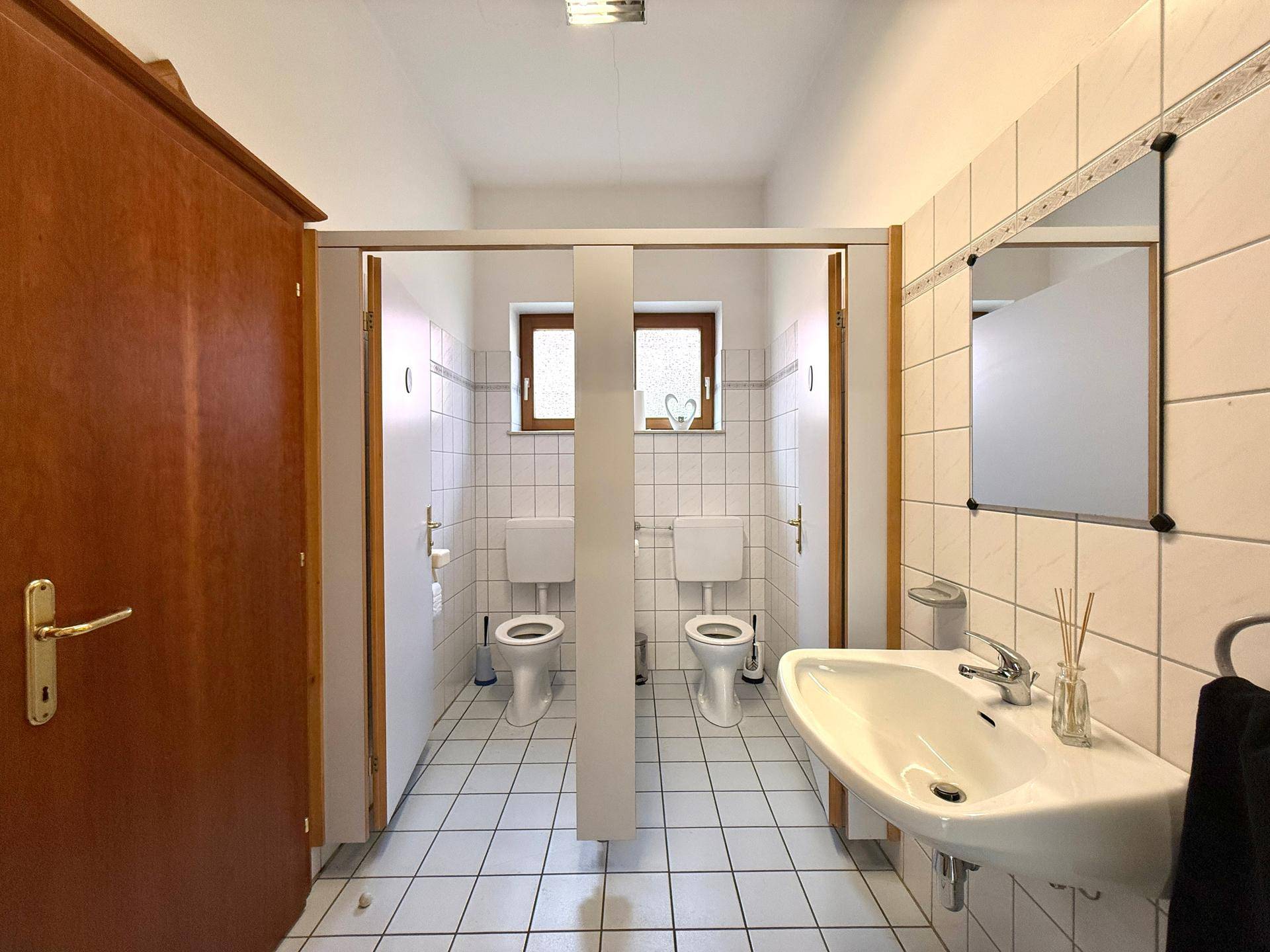 Gefliester Toilettenraum mit zwei Kabinen, Waschbecken und Spiegel, in gutem Zustand.