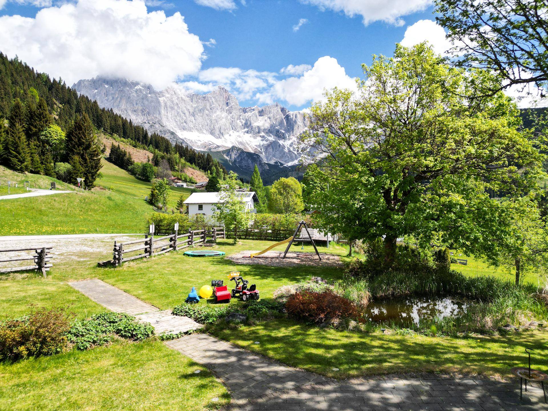 Großer Garten mit Spielplatz, Teich und Bäumen, mit beeindruckendem Bergblick im Hintergrund.