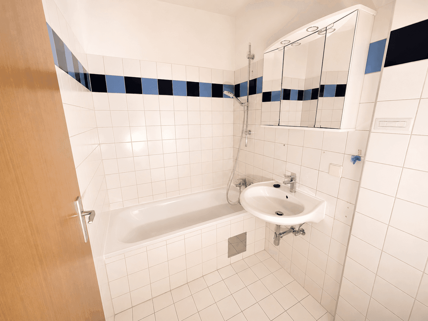 Helles Badezimmer mit Badewanne, Duschfunktion, Waschbecken und Spiegelschrank, akzentuiert durch blaue Fliesen.