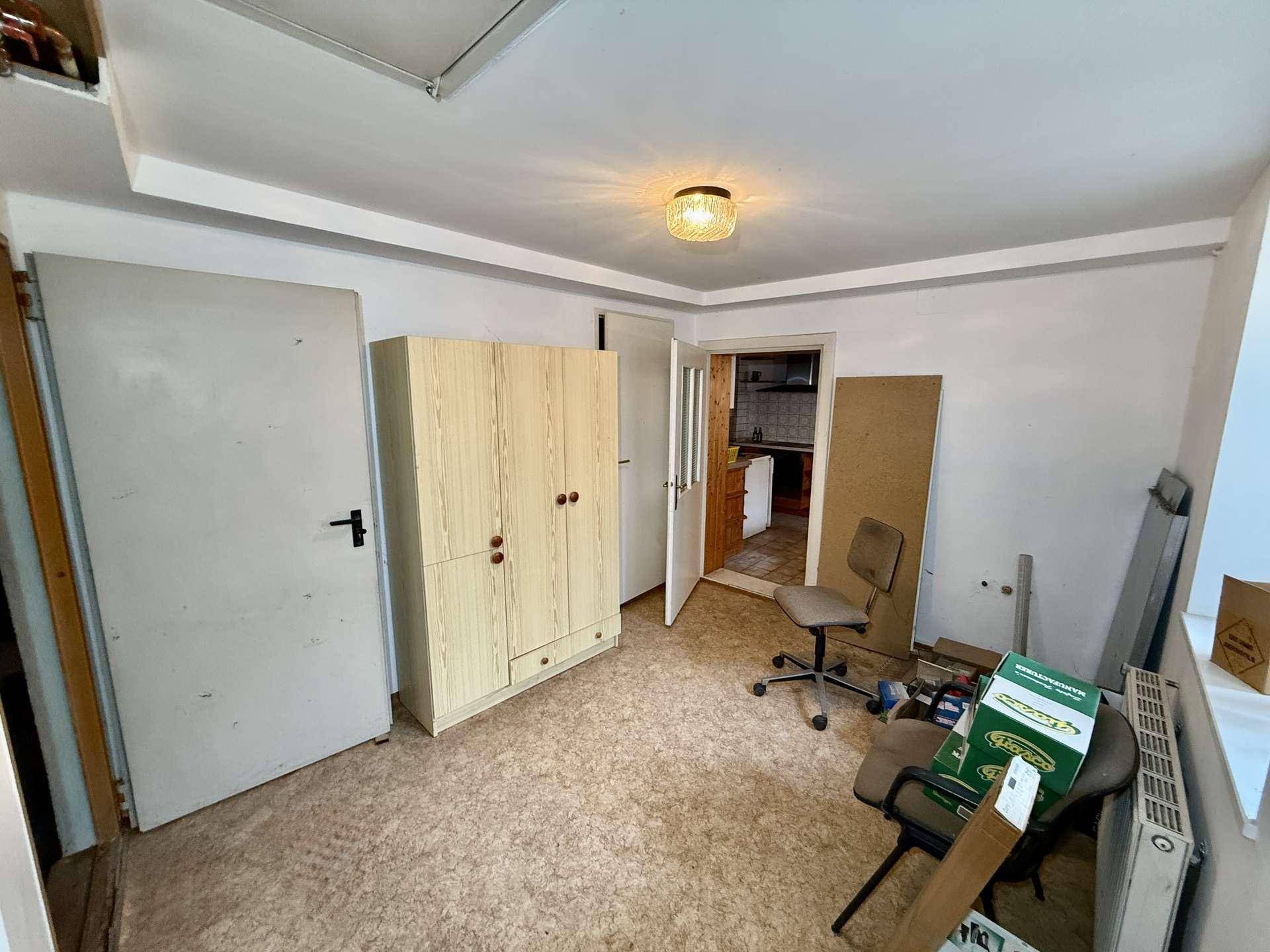 Einfacher Raum mit Linoleumboden, Kleiderschrank und Blick in die angrenzende Küche.