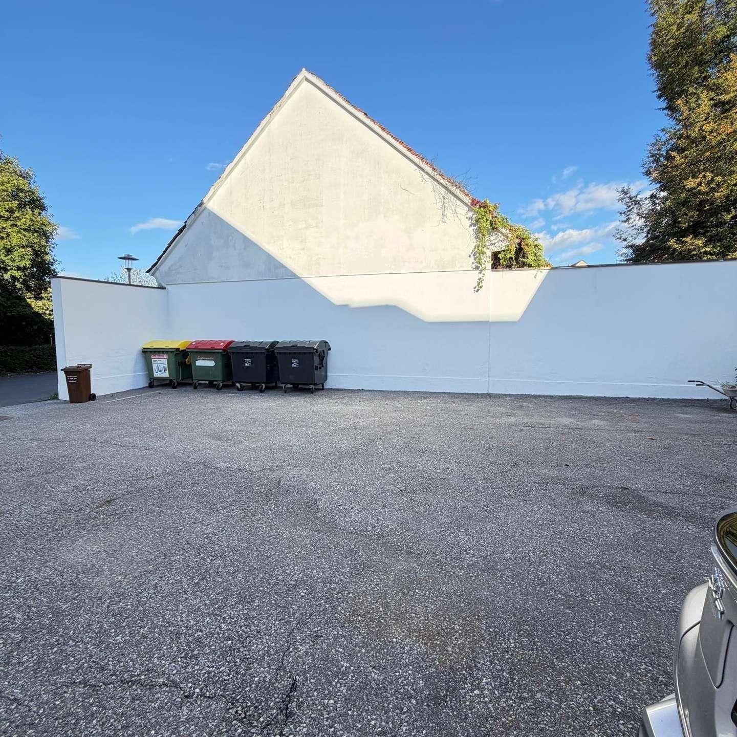Großer Parkplatz mit einer weißen Mauer und Mülltonnen im Hintergrund, umgeben von Bäumen.
