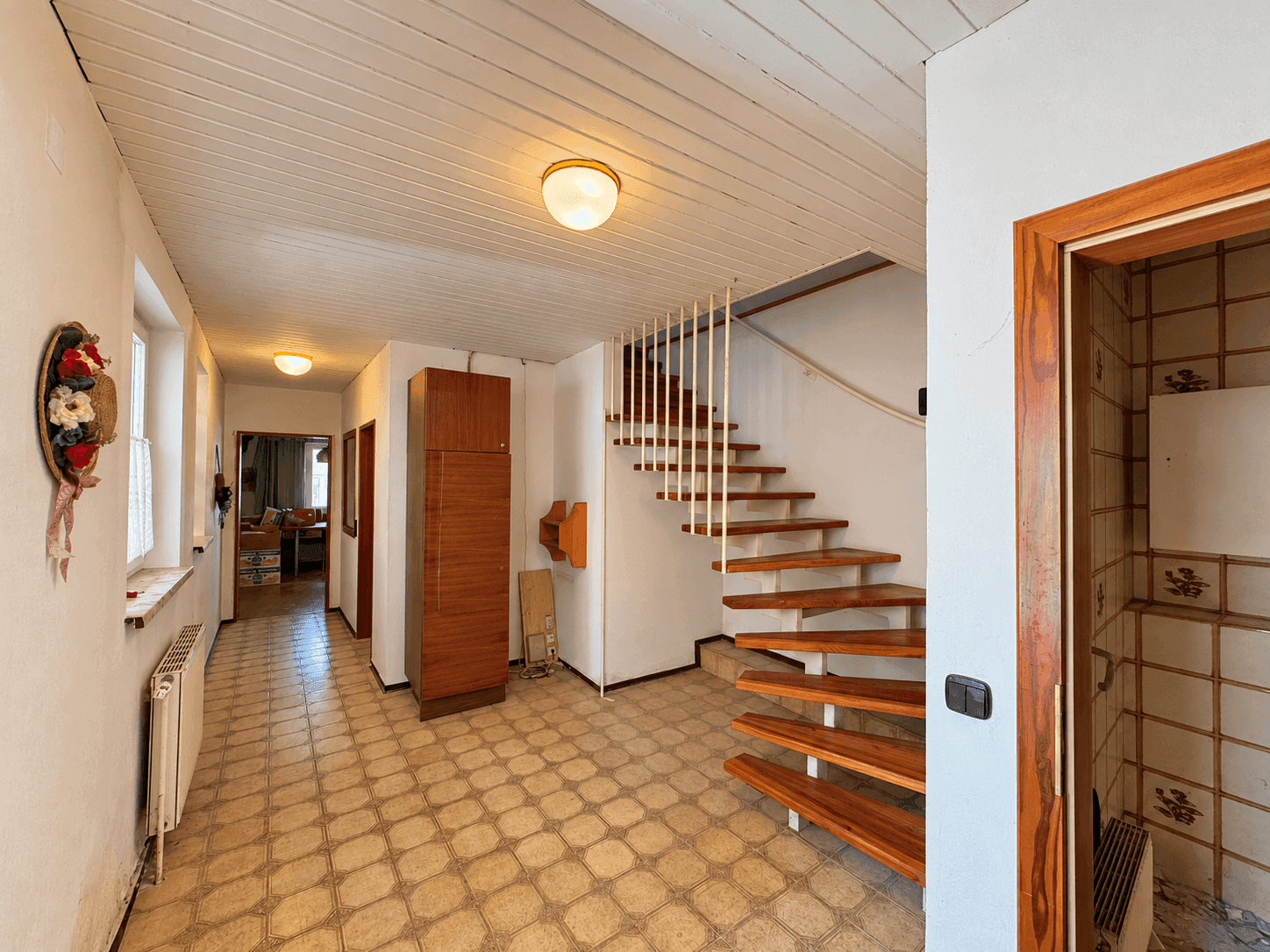 Heller Flur mit Holztreppe, Fliesenboden und Holzpaneelen an der Decke.
