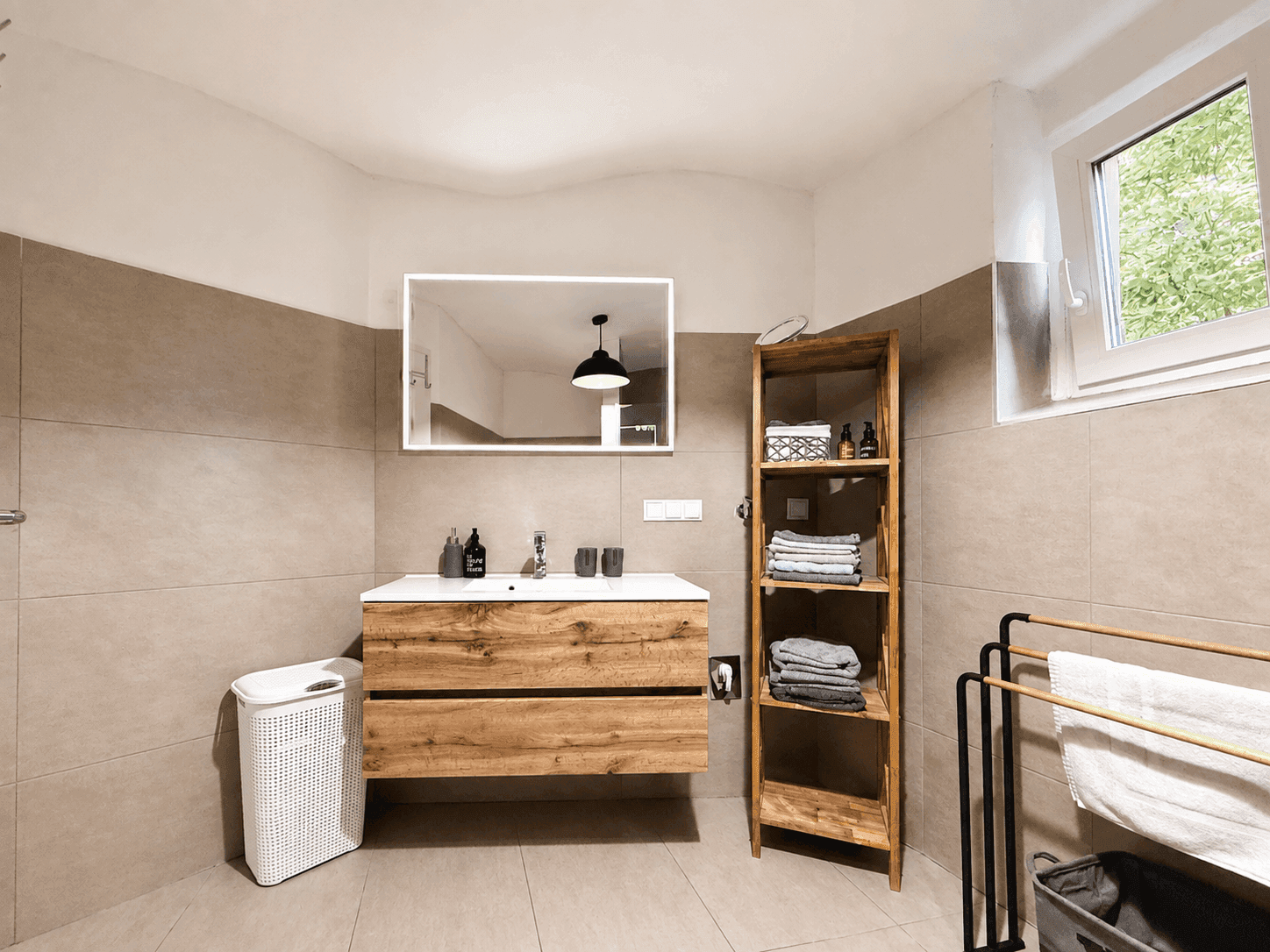 Helles Badezimmer mit modernem Holzwaschtisch, Spiegel und beigefarbenen Wandfliesen.