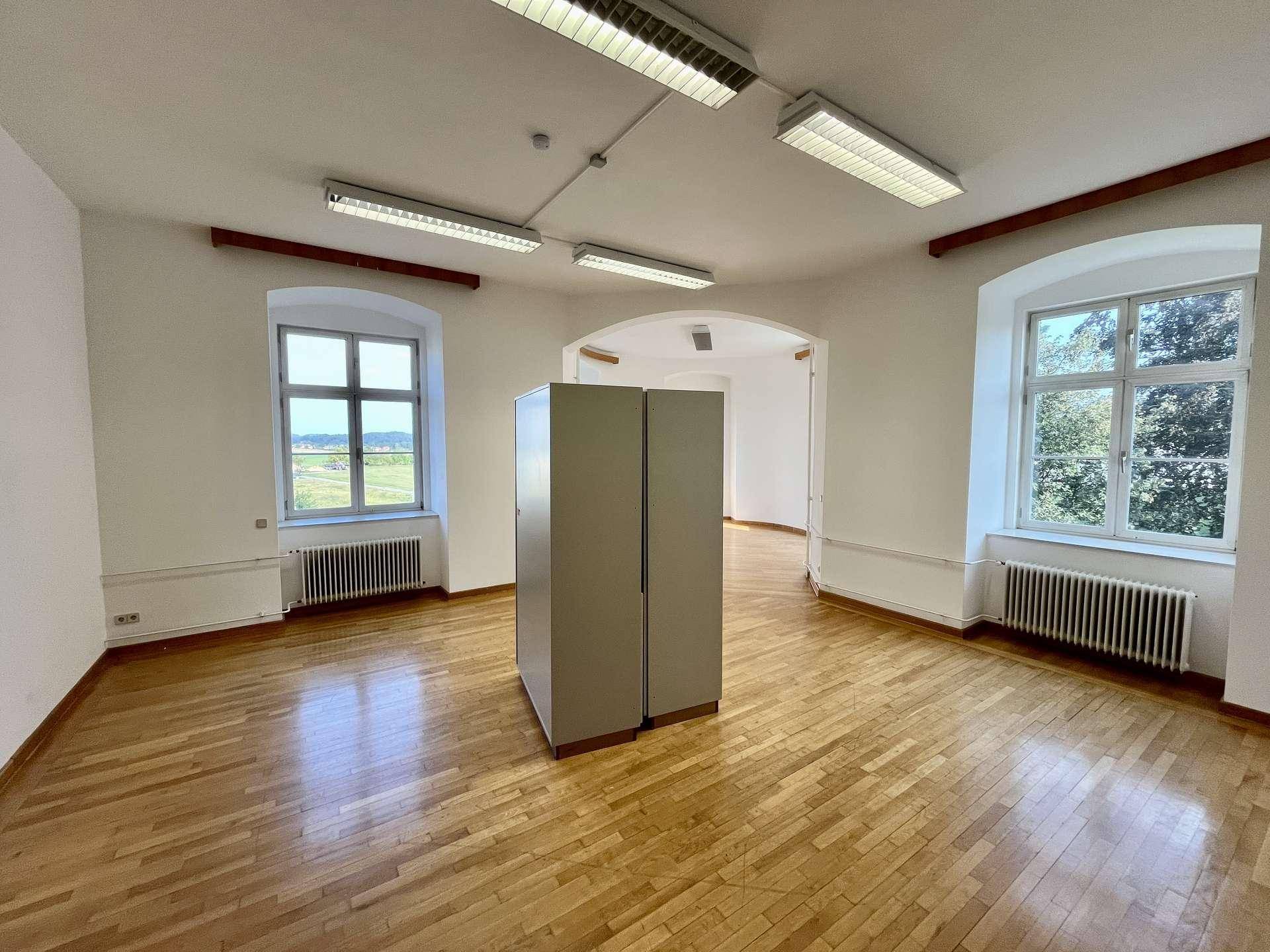 Großer Büroraum mit Holzboden, zwei Fenstern und einem Schrank in der Mitte des Raumes.
