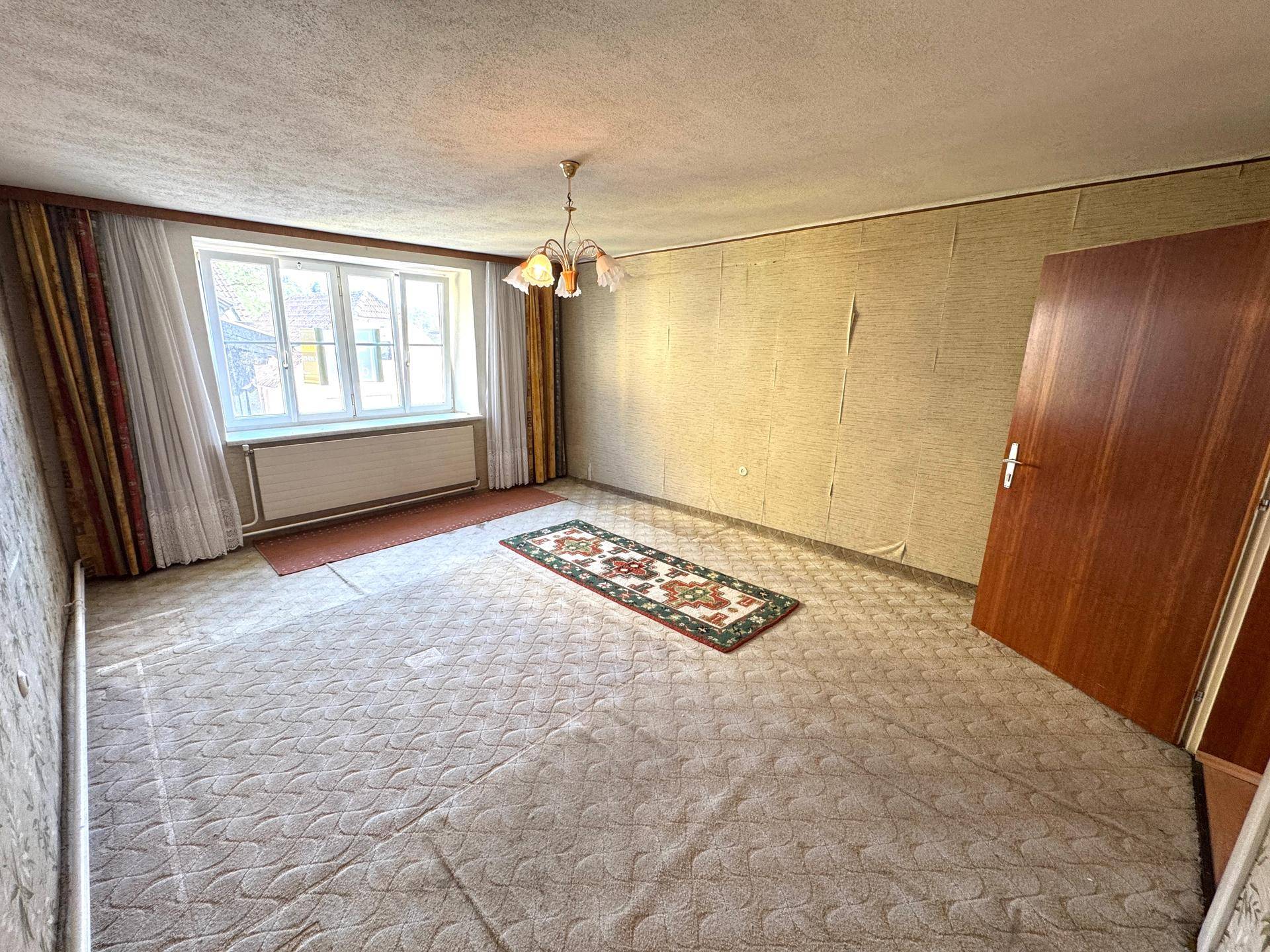 Schlafzimmer mit Teppichboden, Tapetenresten und Blick auf die Umgebung.