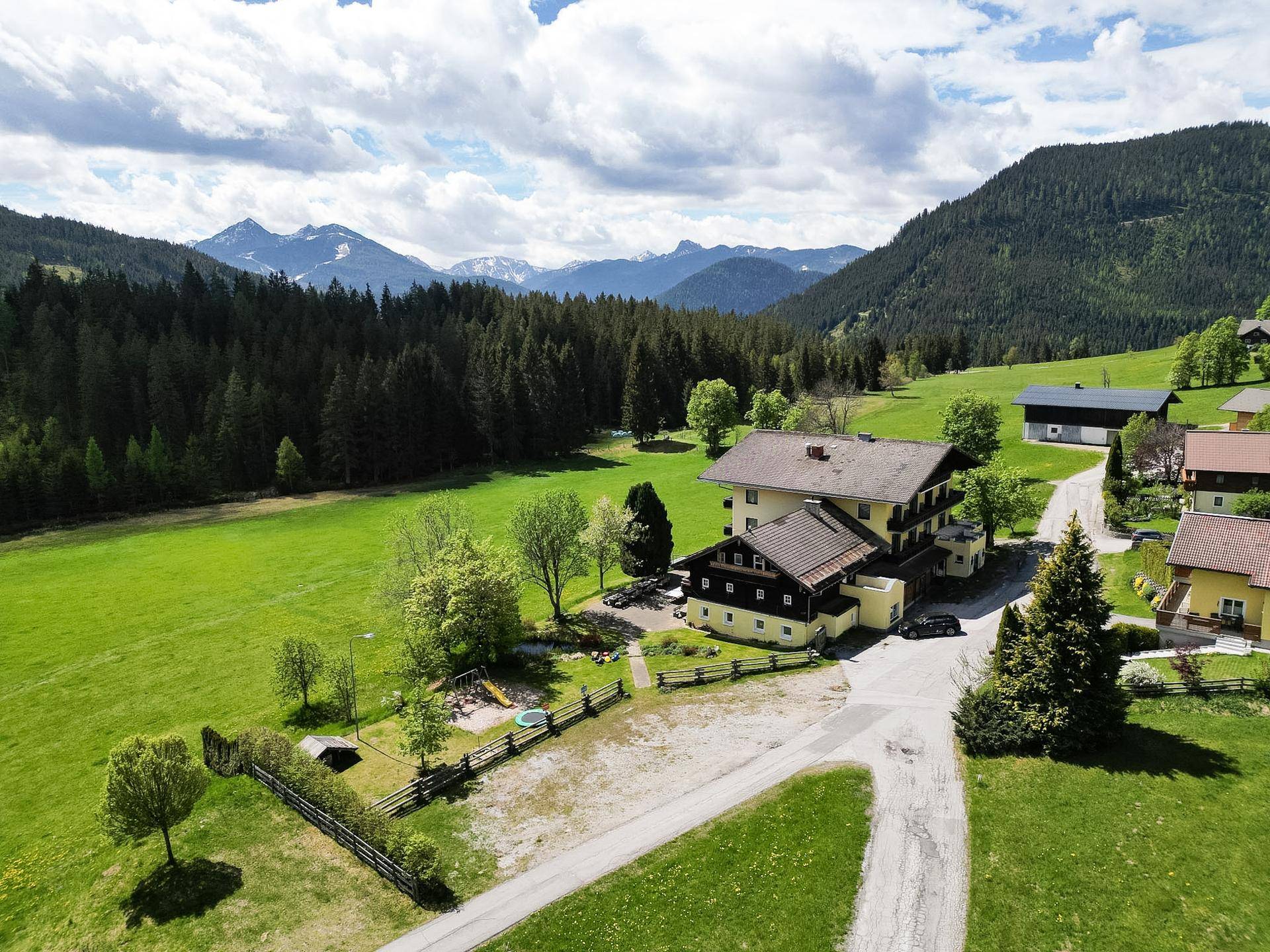 Luftaufnahme eines Anwesens mit mehreren Gebäuden, umgeben von Wiesen, Wäldern und einer Bergkulisse.