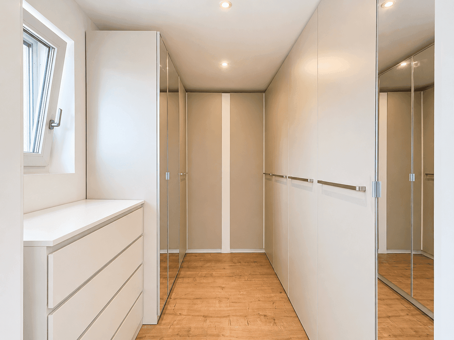 Geräumiger begehbarer Kleiderschrank mit Einbauschränken, Spiegeln und Fenster für Tageslicht.