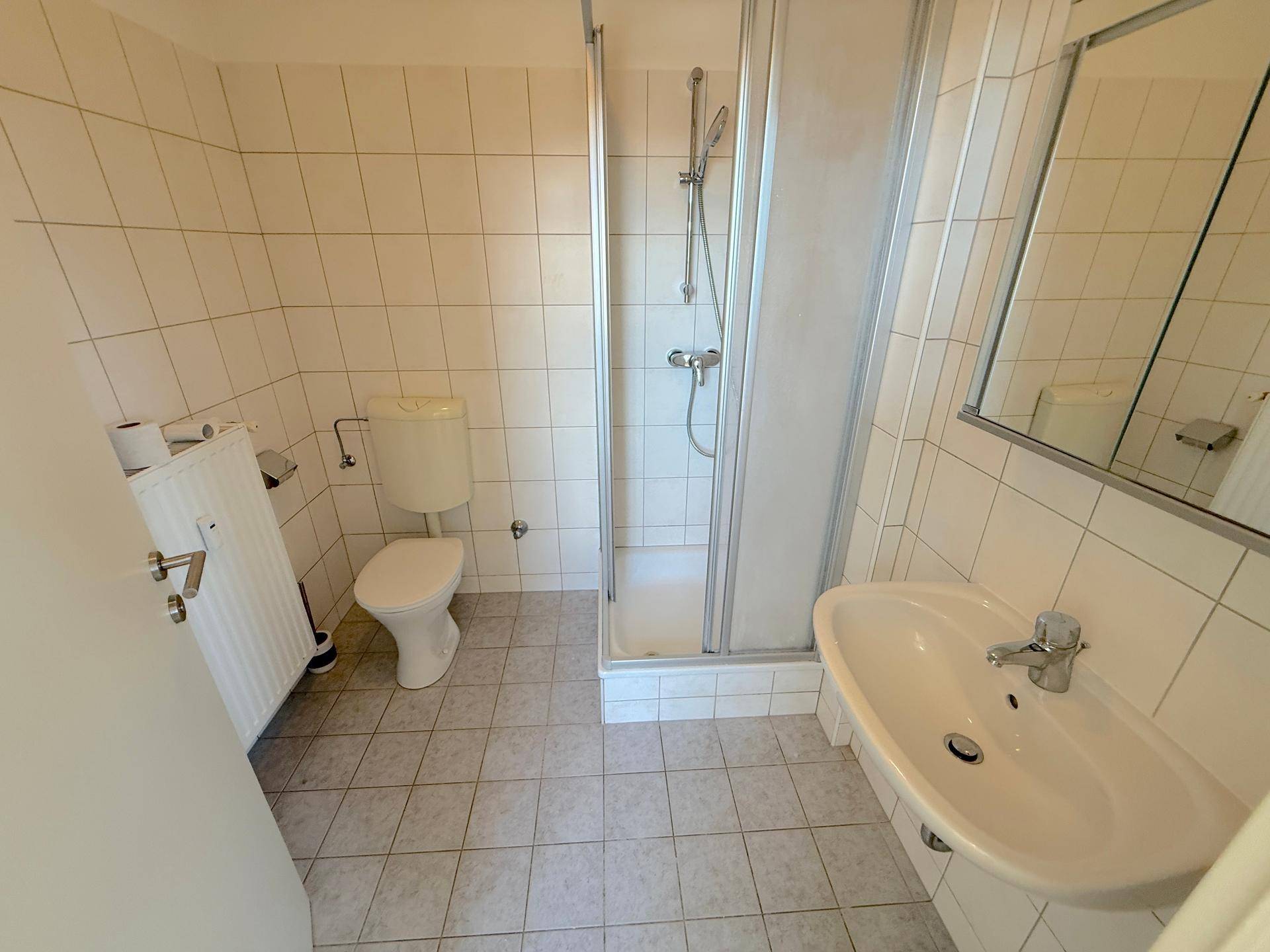 Funktionales Badezimmer mit weißen Fliesen, Dusche, WC, Waschbecken und Heizkörper.