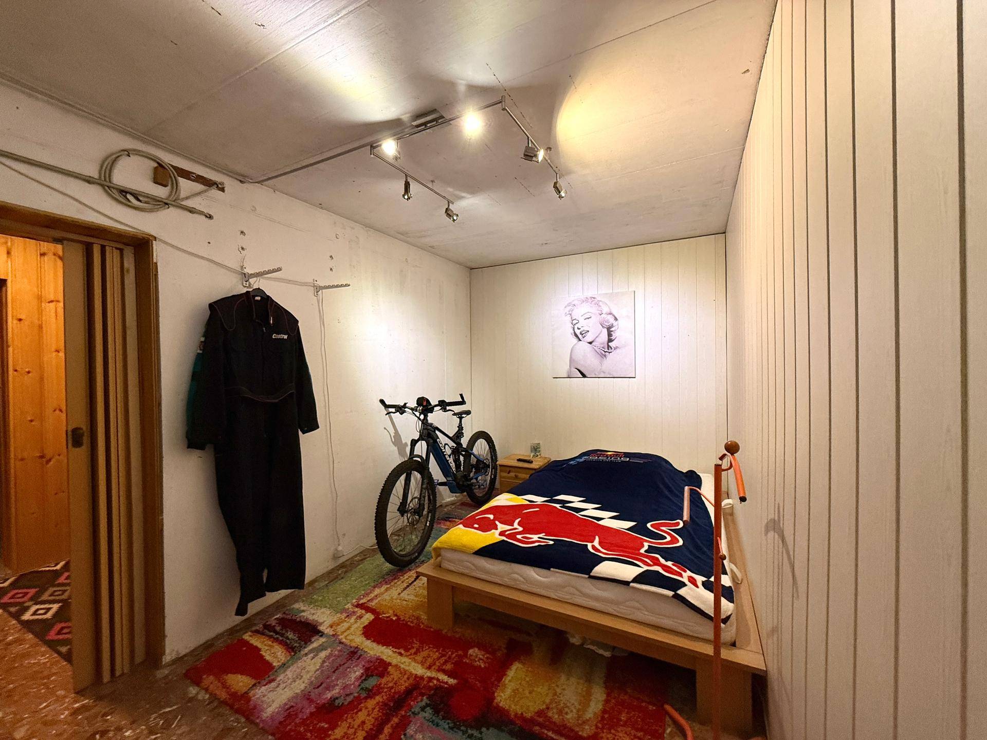 Einfaches Schlafzimmer im Untergeschoss mit Bett, Fahrrad und Marilyn Monroe Bild.