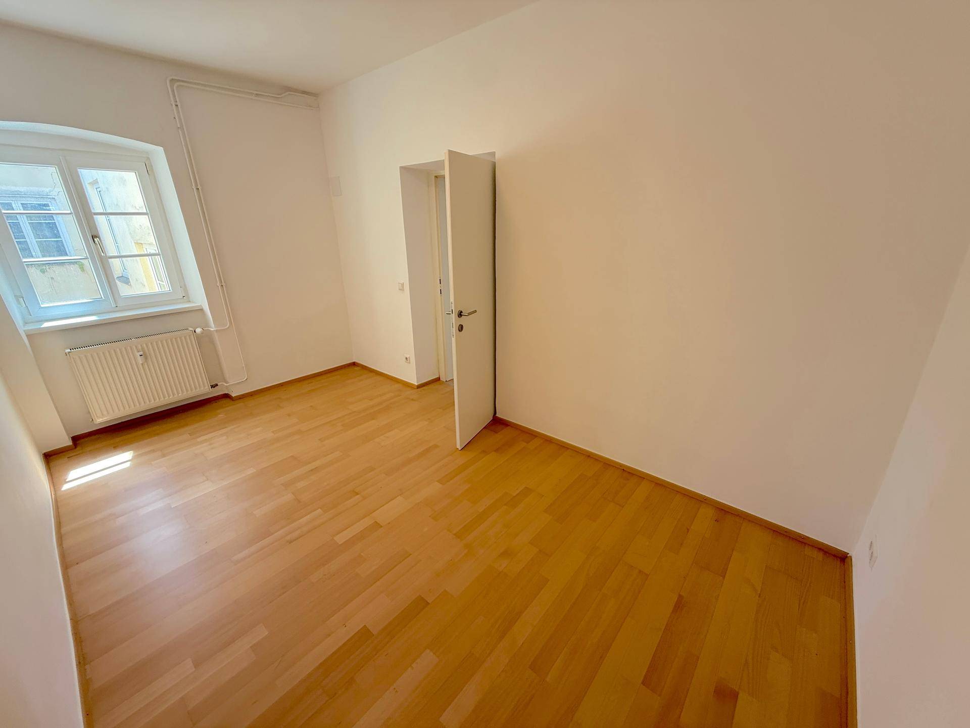 Helles Schlafzimmer mit Holzboden, weiß gestrichenen Wänden und einem Fenster mit Heizkörper.