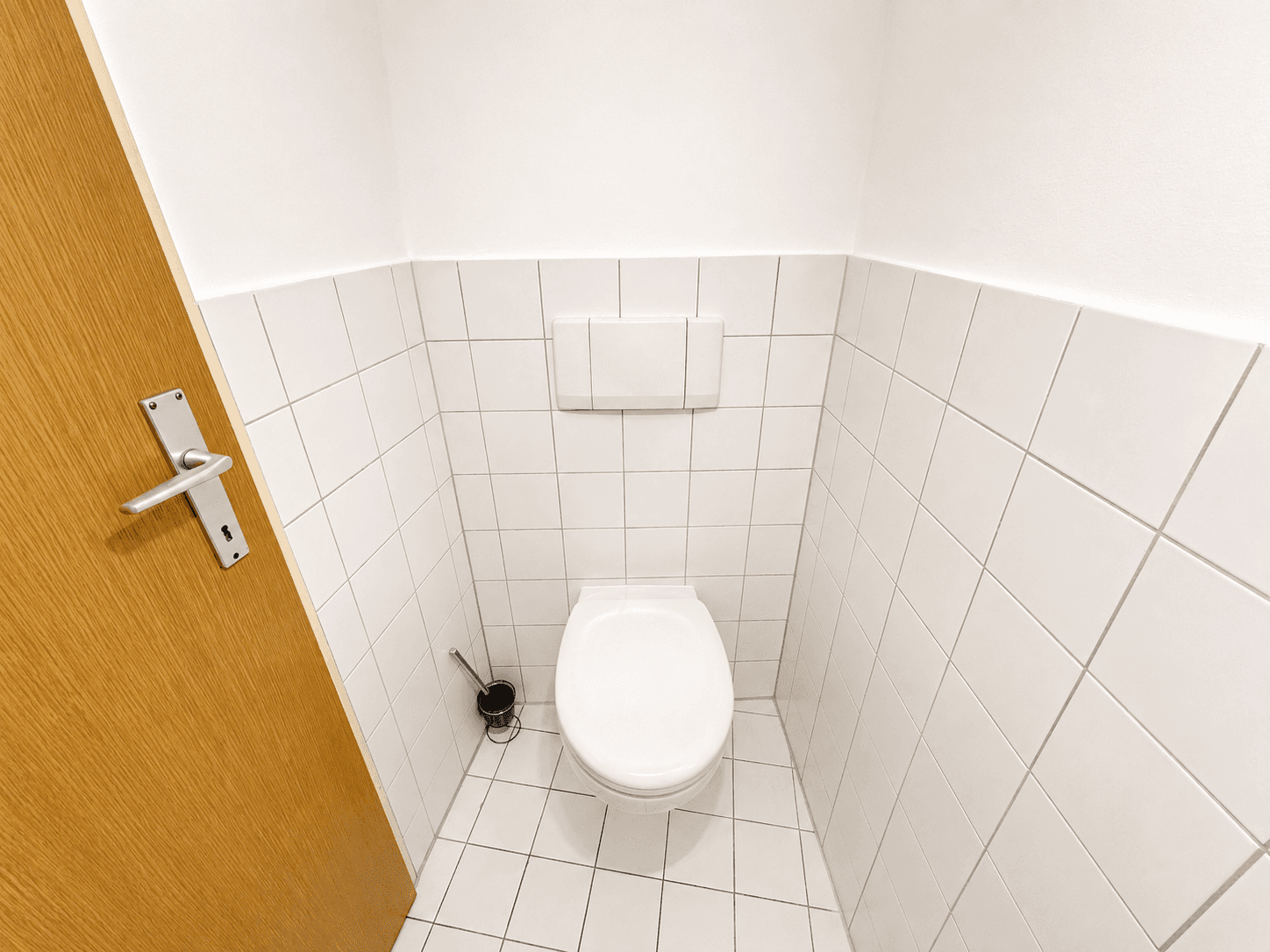 Separate Toilette mit wandmontiertem WC und weißen Fliesen an Wänden und Boden.