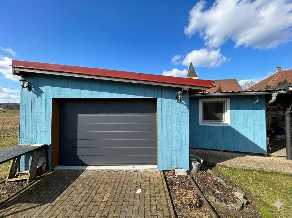 Blaue Holzgarage mit modernem Garagentor und gepflasterter Zufahrt.