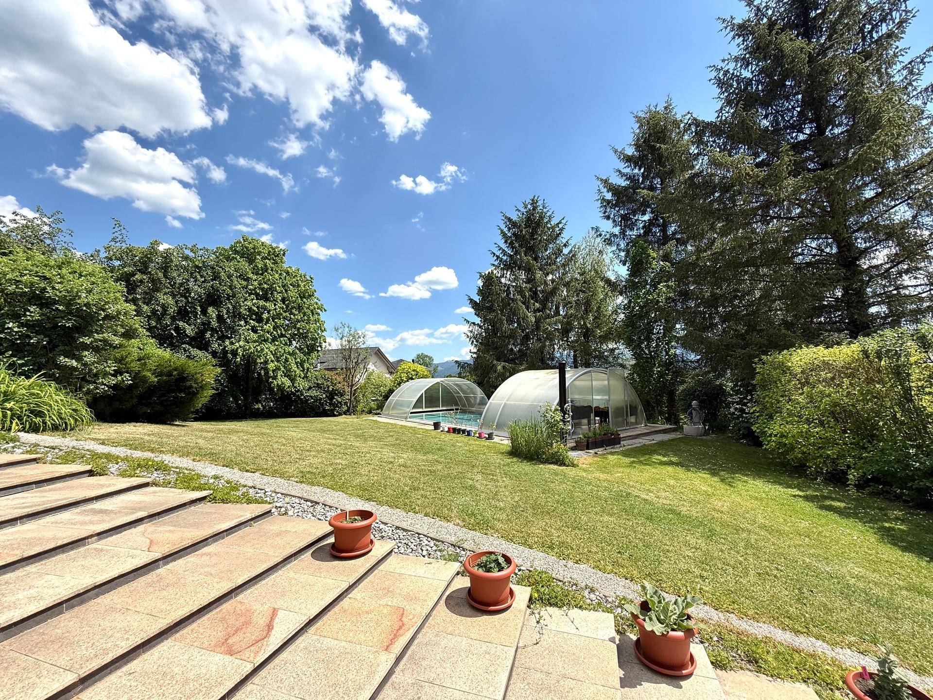 Großer Gartenbereich mit Rasenfläche und zwei überdachten Swimmingpools im Hintergrund.